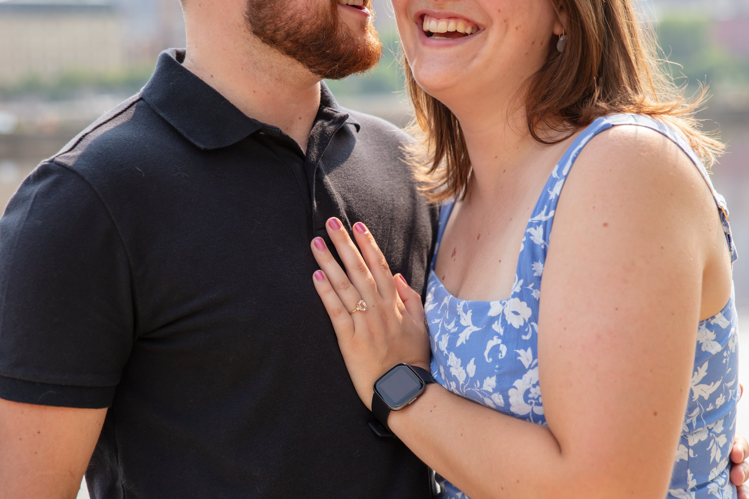 Close up image of a couple, a man and woman, embracing each other and smiling at the camera after he proposed to her in front of the falls and skyline at Water Power Park on St. Anthony Main. The photo focuses on her left hand on his chest showing of