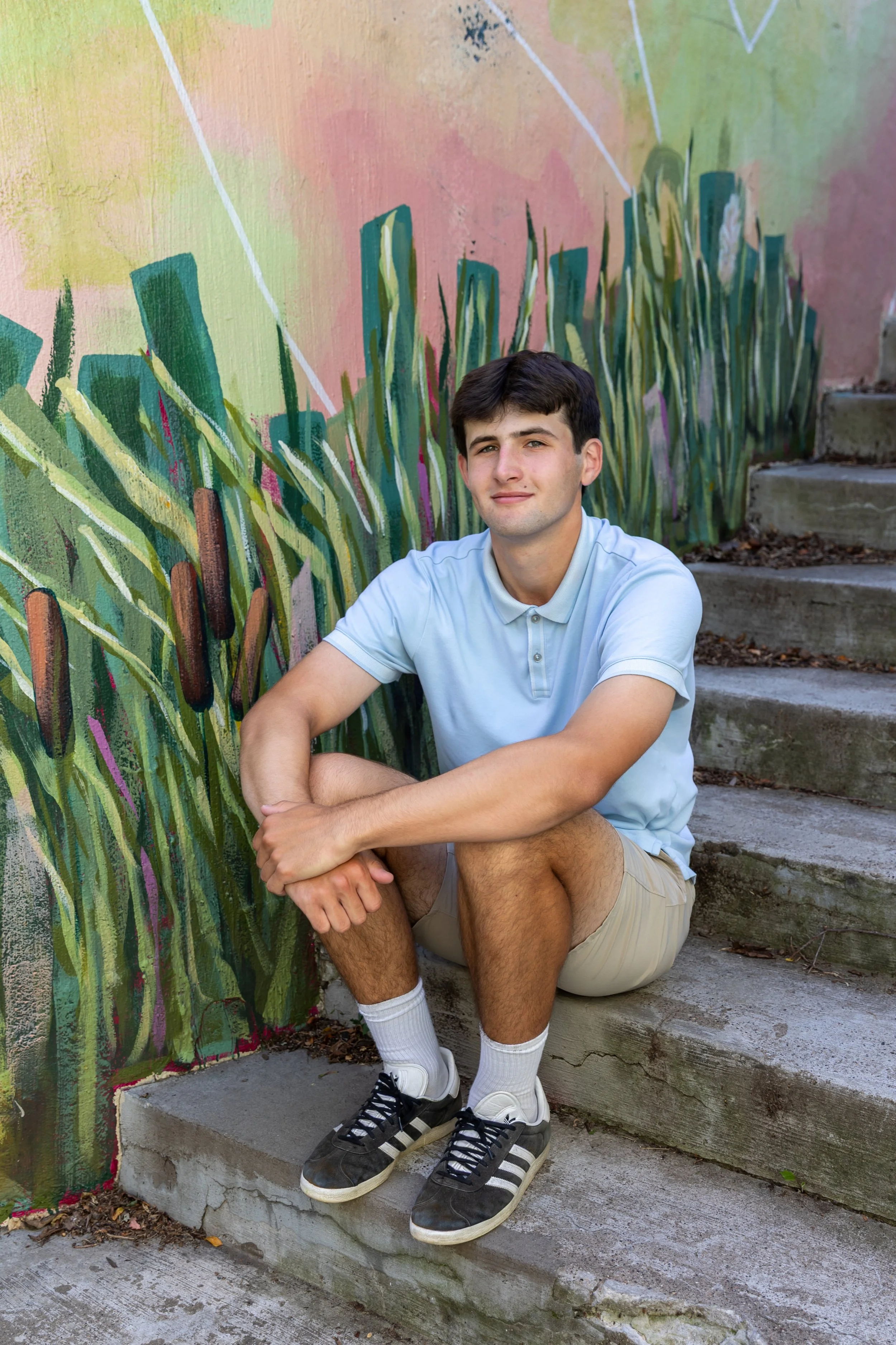 A Minneapolis high school sits on some steps with their arms crossed on their knees in front of a mural at Lake Harriet smiling at the camera for their senior photos.