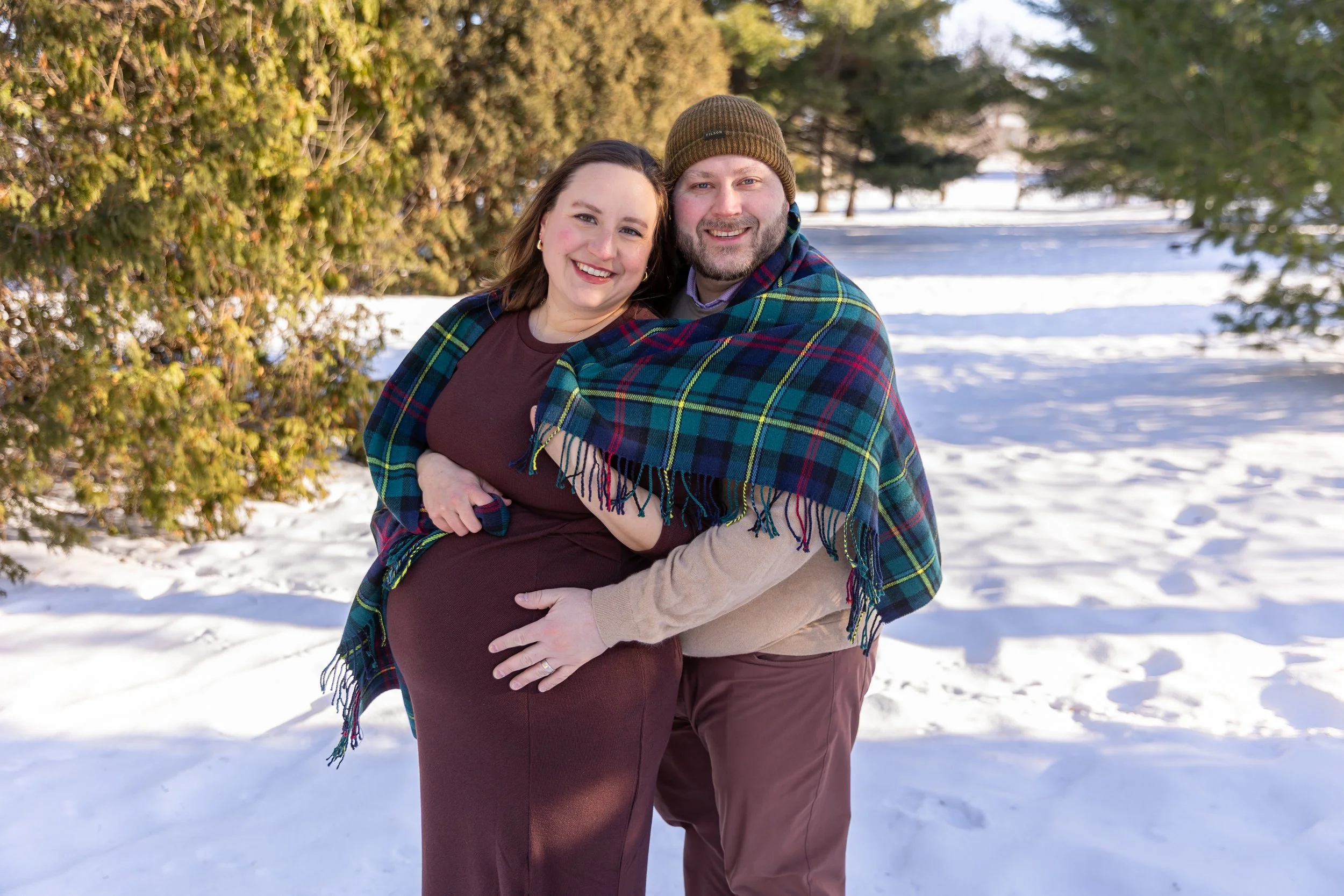 Maternity session, expecting mother and father-to-be embrace each other smiling at the camera while wearing a blanket at Arneson Acres Park in Edina, MN in the winter.
