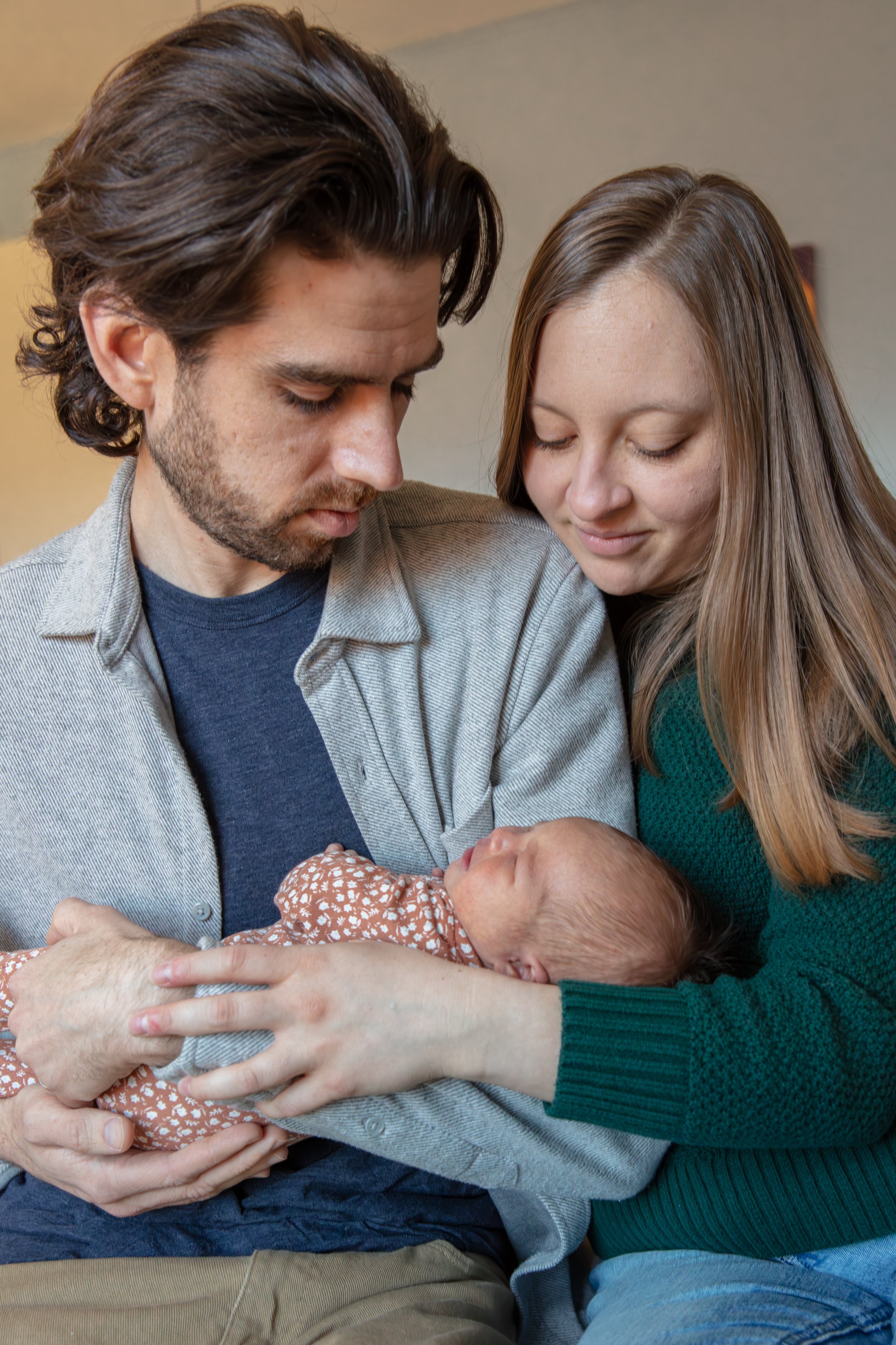 Newborn in-home session - father and mother hug together smiling down as the father holds the newborn in his arms.