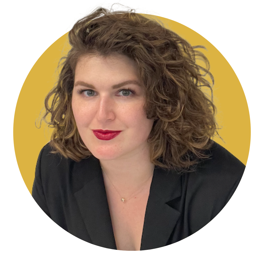 A woman with curly, shoulder-length brown hair wearing a black blazer and red lipstick, against a mustard yellow background.