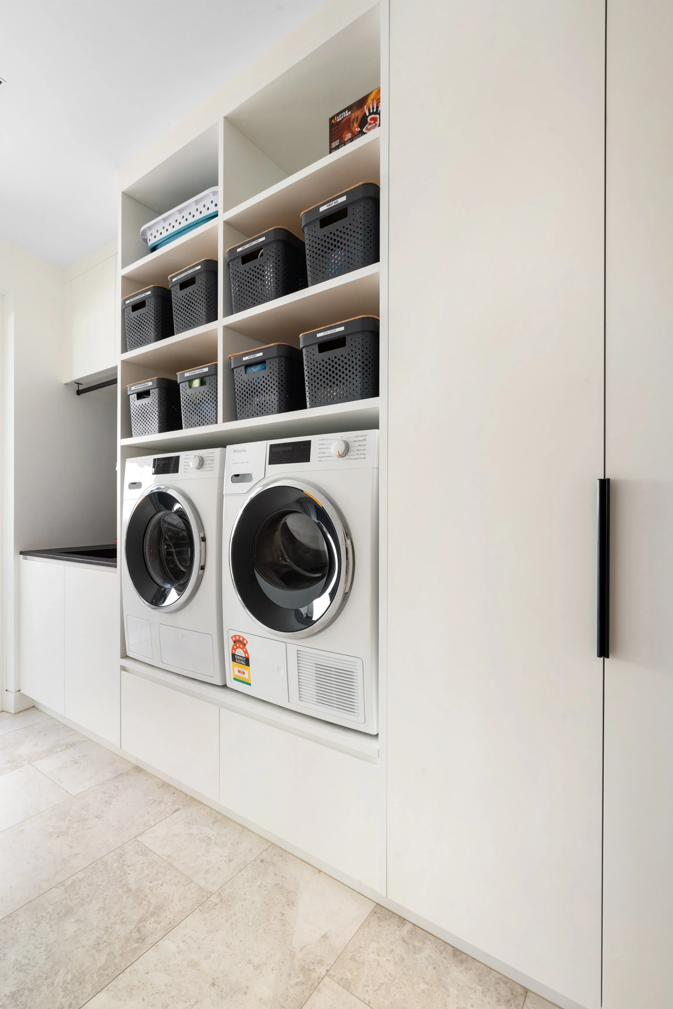 Sleek laundry room with a front-loading washing machine and dryer, white shelving with black laundry baskets, and built-in cabinets.