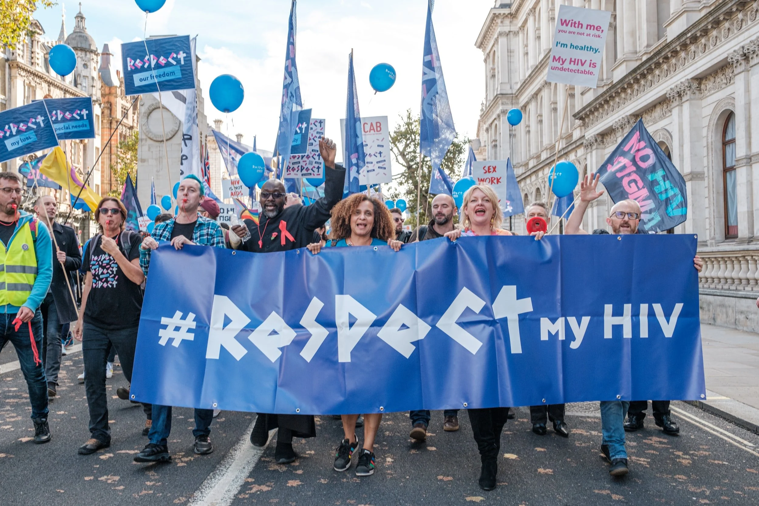 Group of people marching with blue banner reading '#Respect my HIV' at a protest. Participants hold signs and balloons, advocating for HIV awareness and rights.
