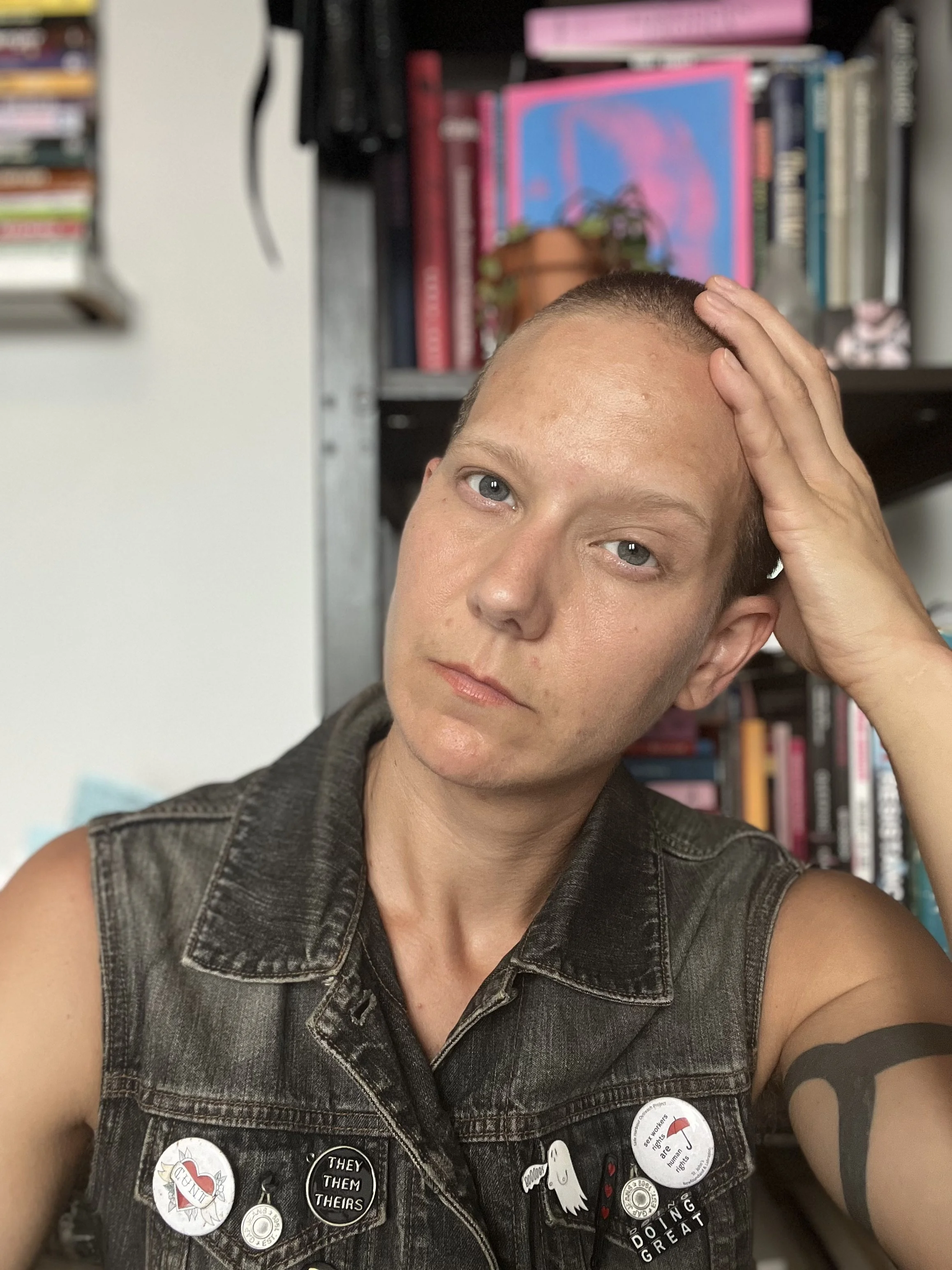 Person with short hair wearing a denim vest adorned with pins, posing indoors in front of a bookshelf.