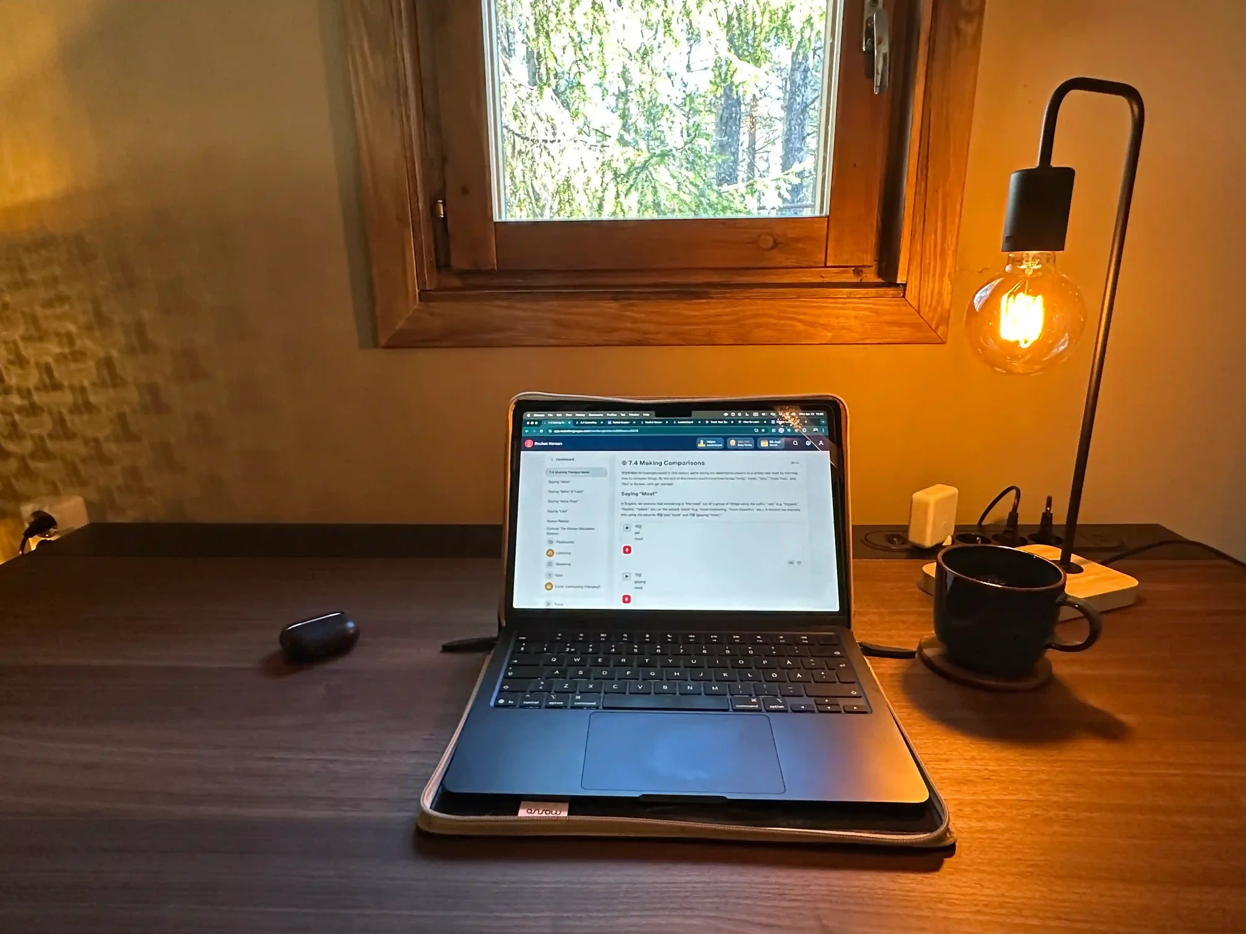 A warmly lit home office at early morning with a MacBook open to a Rocket Korean lesson, a black coffee mug, and an Edison bulb table lamp casting amber light across a walnut desk.