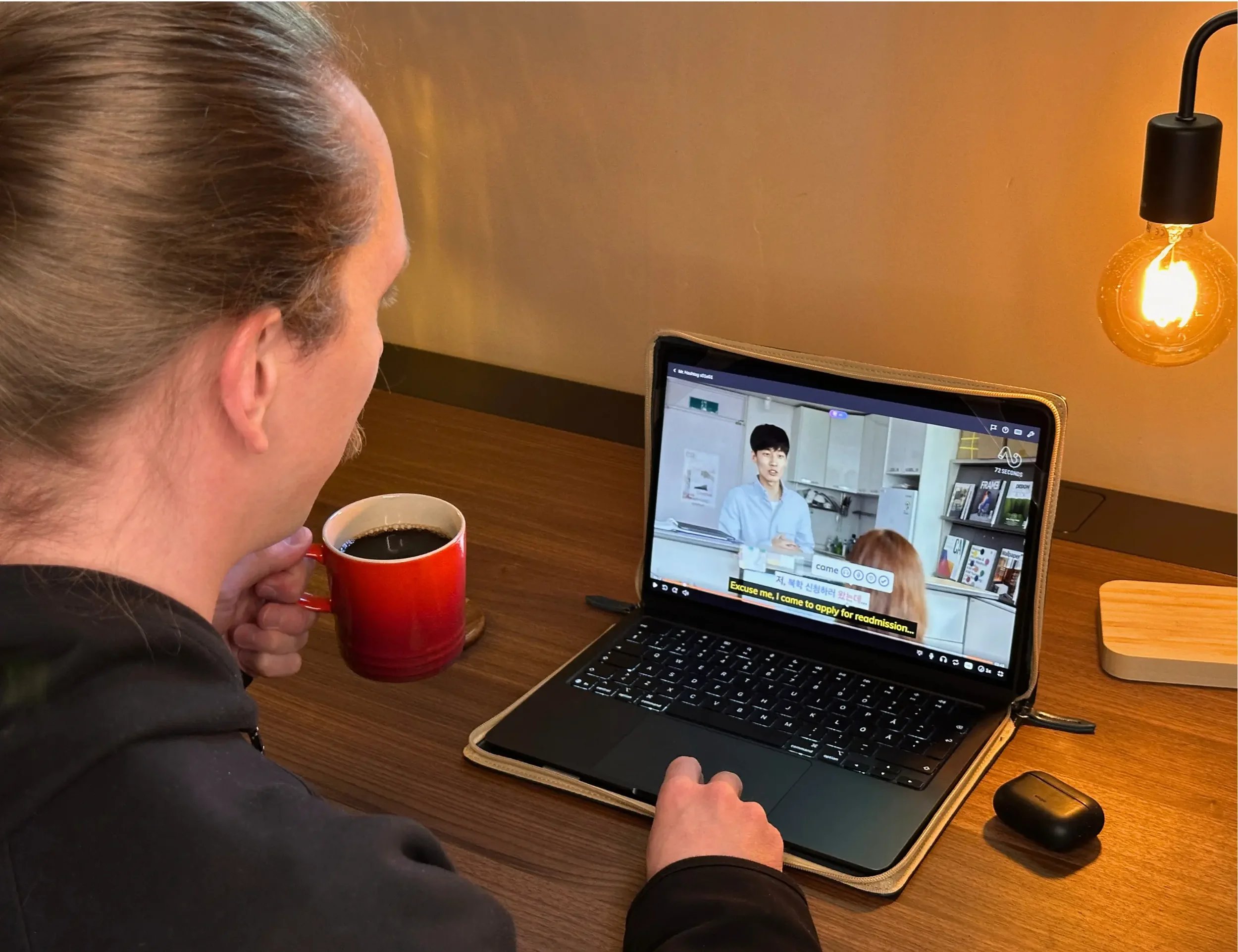 Over-the-shoulder view of a language learner watching a Korean show on Lingopie with the word "came" being saved, holding black coffee in an orange mug at a walnut desk lit by an Edison bulb.