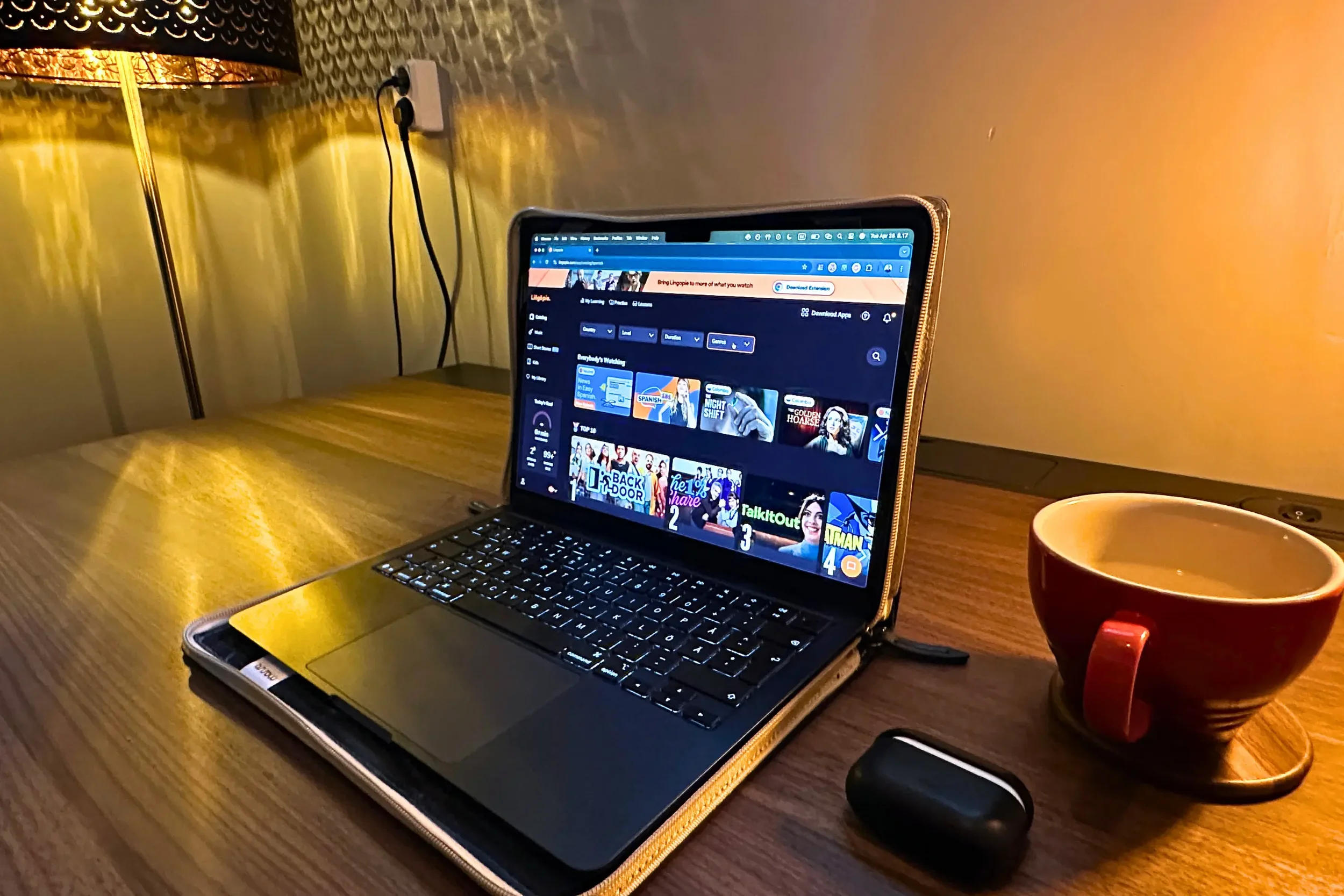 A laptop showing Lingopie's Spanish content catalog, with a red coffee mug, AirPods case, and warm amber lamp lighting in a cozy home office setting.