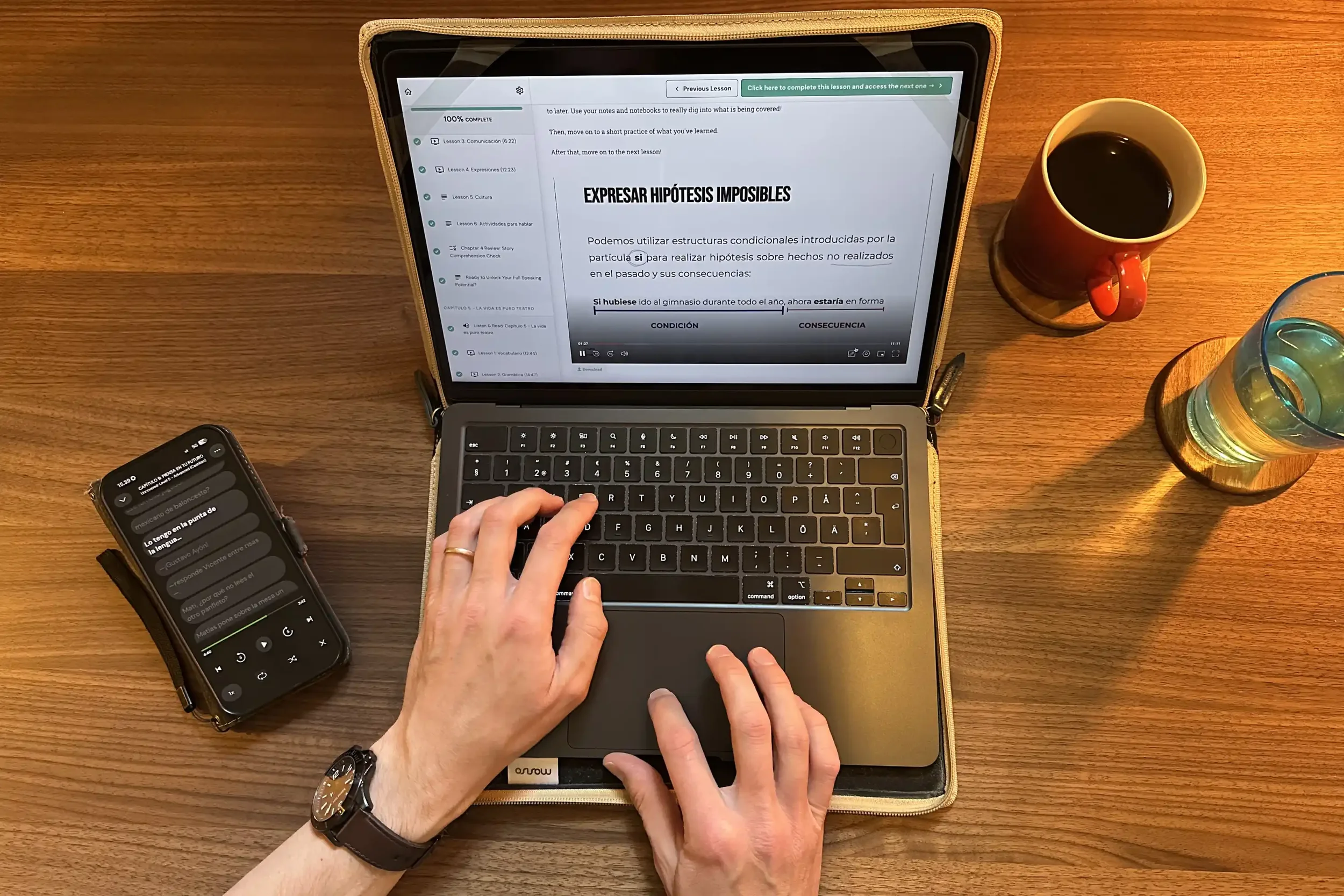 A laptop on a walnut desk showing a Spanish Uncovered Advanced grammar lesson taught in Spanish, titled "Expresar hipótesis imposibles," with a phone beside it displaying the story transcript in LingQ, a mug of coffee and a glass of water nearby.