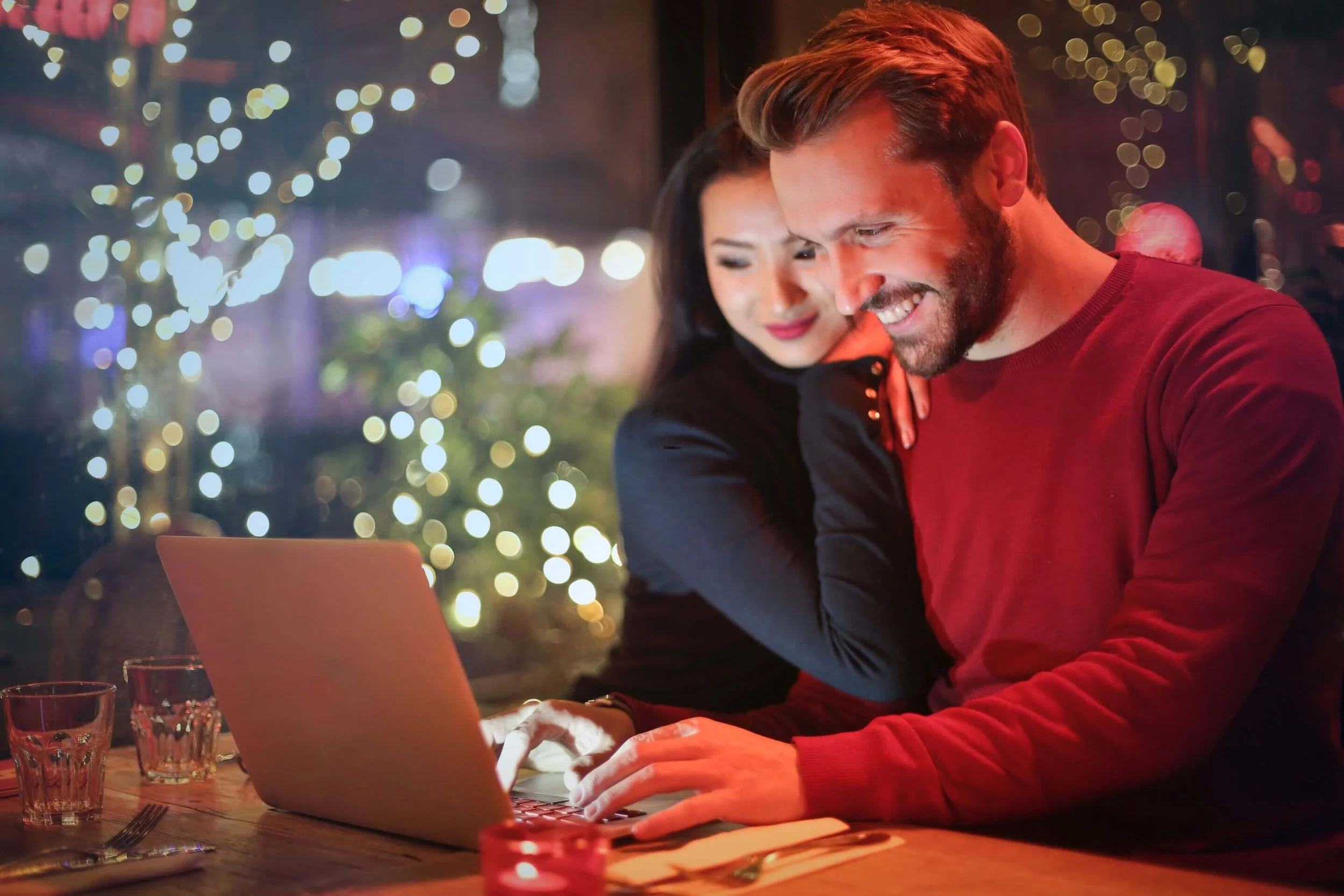 Festive cafe scene with smiling couple huddled over laptop amid twinkling holiday lights