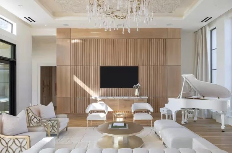 Modern living room with white and beige furniture, a white grand piano, a large flat-screen TV on a wooden accent wall, a chandelier, and large windows with curtains.