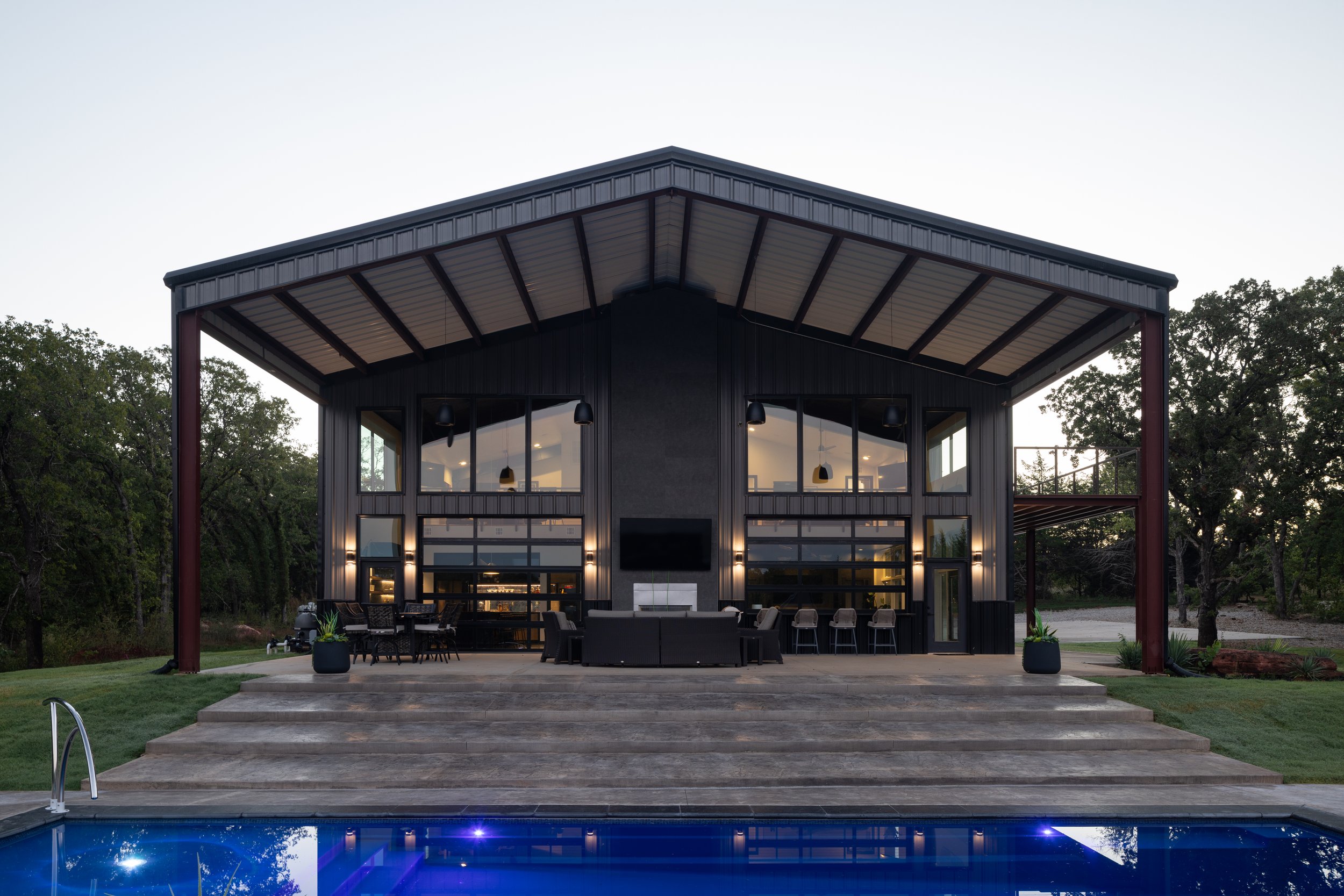 Modern two-story house with large windows and an outdoor patio area, surrounded by trees and greenery, with a swimming pool in the foreground at dusk.