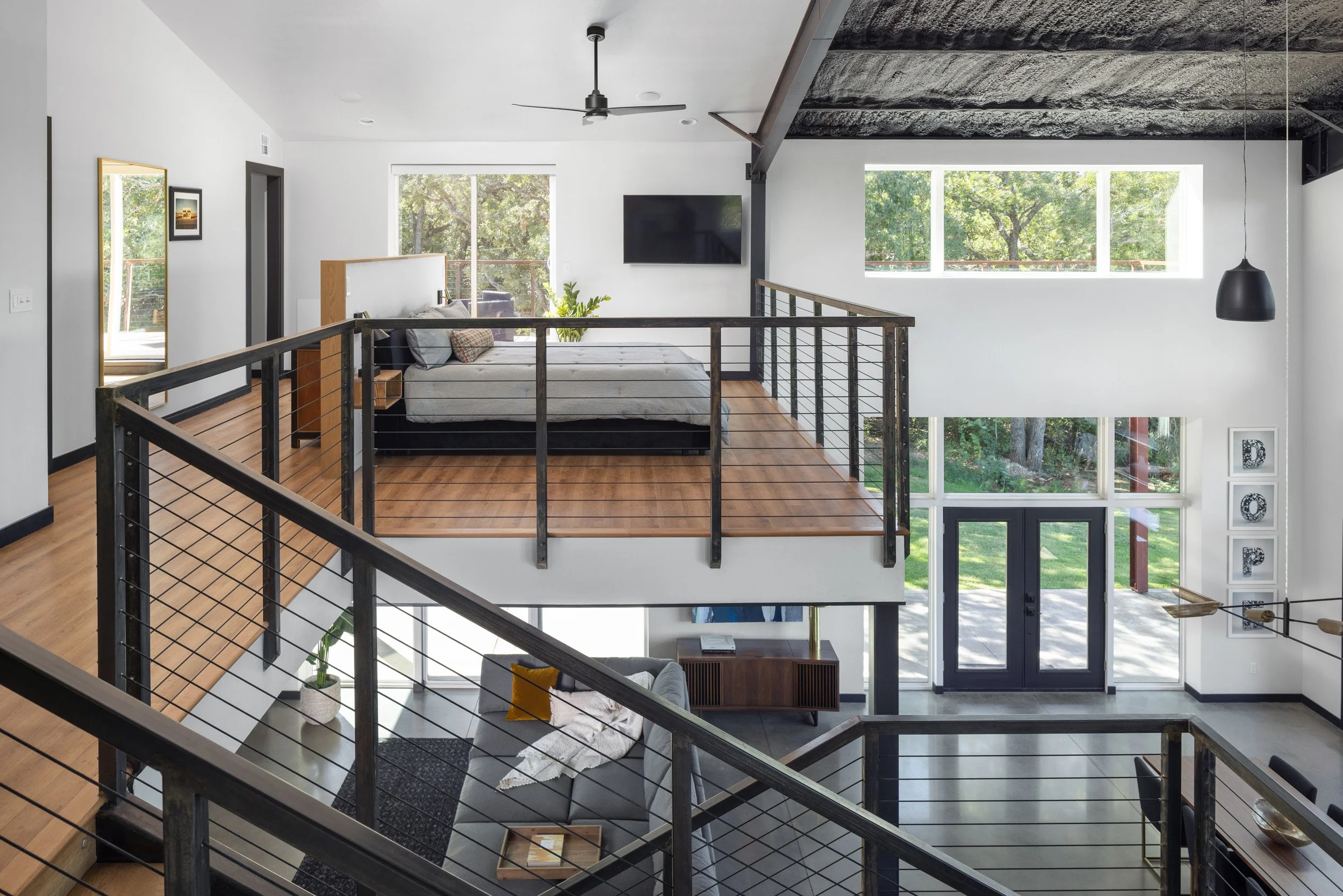 Interior of a modern, open-concept home with a loft area, featuring a black metal railing, wooden floors, a bed with gray bedding, large windows with trees outside, and a black wall-mounted TV.