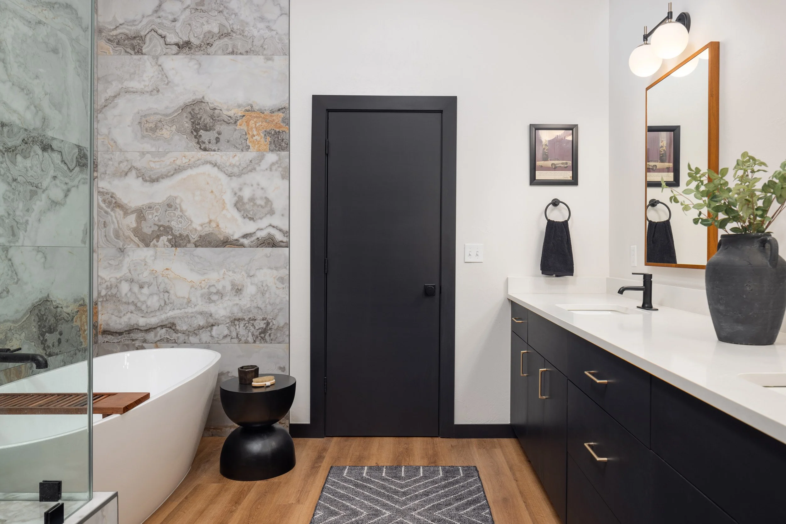 Modern bathroom with a black door, marble wall, white bathtub, black side table, black towel, vanity with black cabinets, white countertop, large mirror, framed picture, potted plant, and ceiling lights.