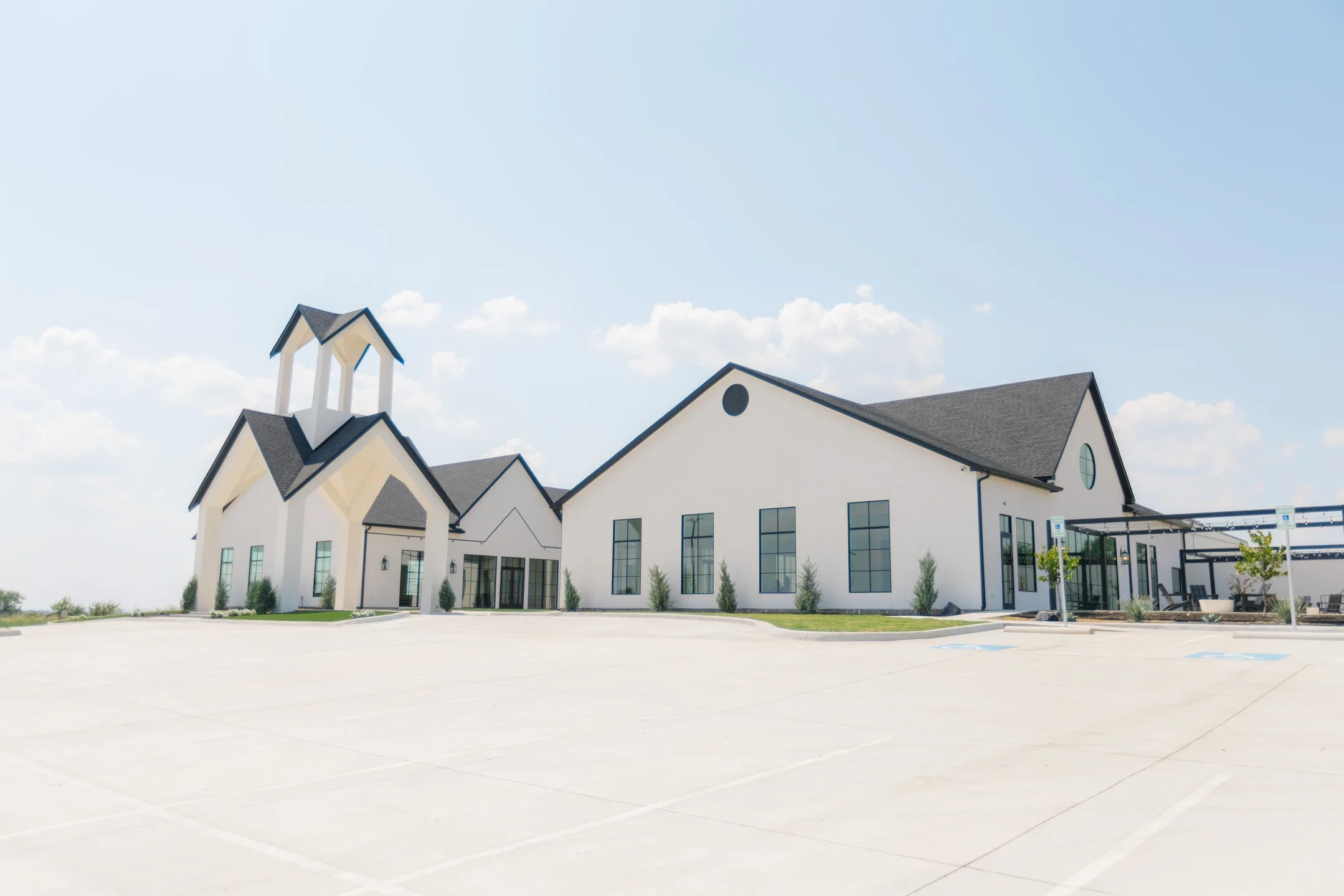 White church-like building with a steeple, large windows, and a parking lot in front, under a clear blue sky.