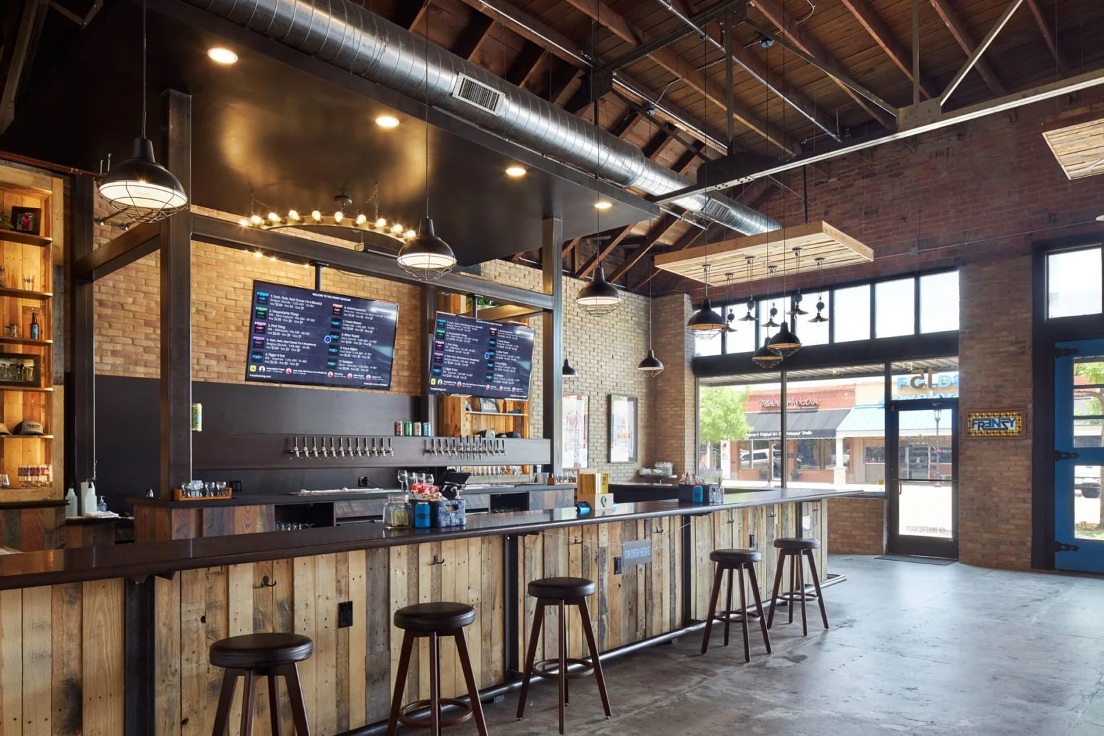 Empty bar with a wooden counter and stools, two digital menu screens hanging above, exposed brick walls, large windows letting in natural light, ceiling with visible ductwork and pendant lights.