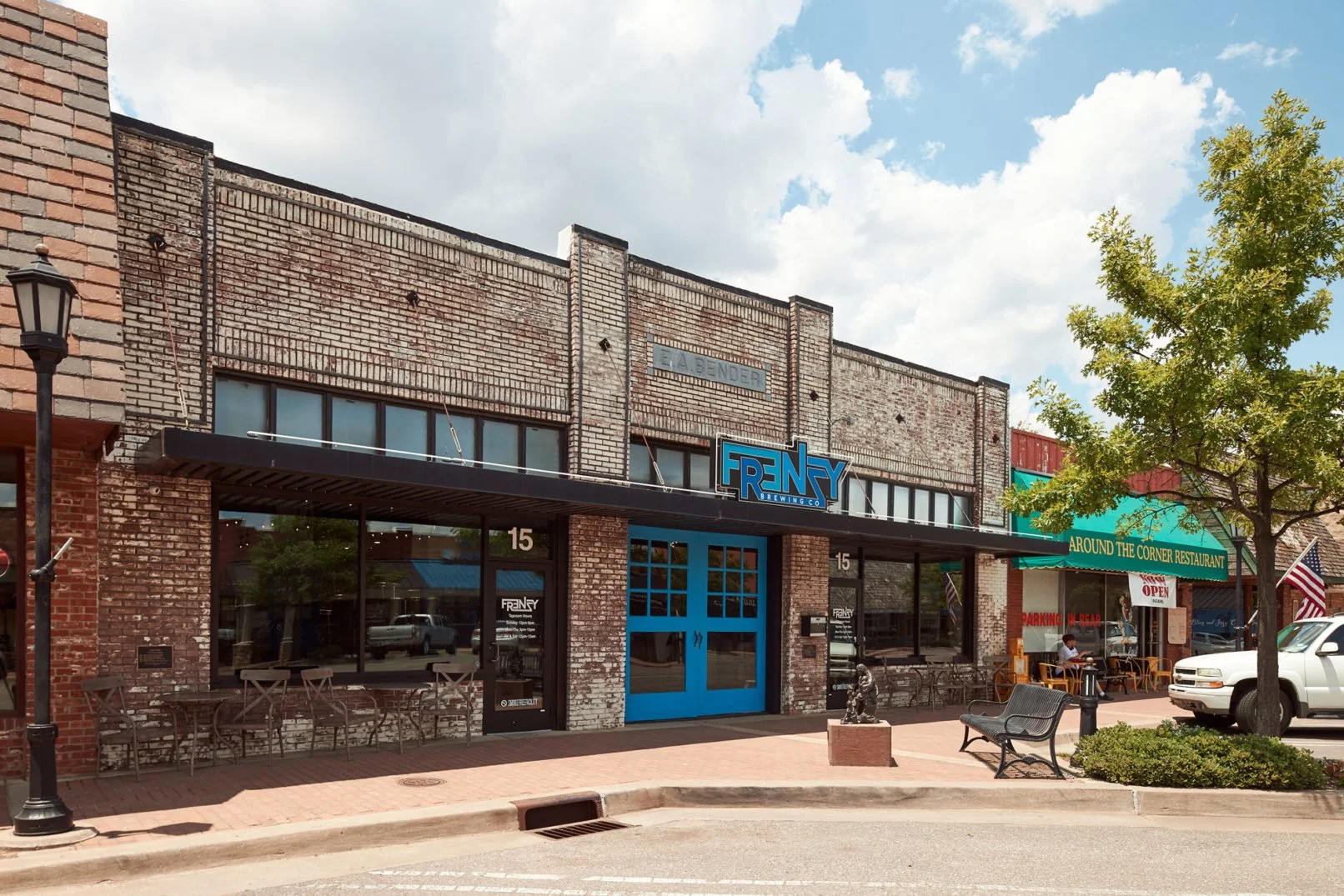 Street view of a brick building with a businesses sign that says 'Freny Brewing Co' and outdoor seating, a restaurant with a green awning reading 'Around the Corner Restaurant,' trees, parked cars, and a sidewalk with benches.