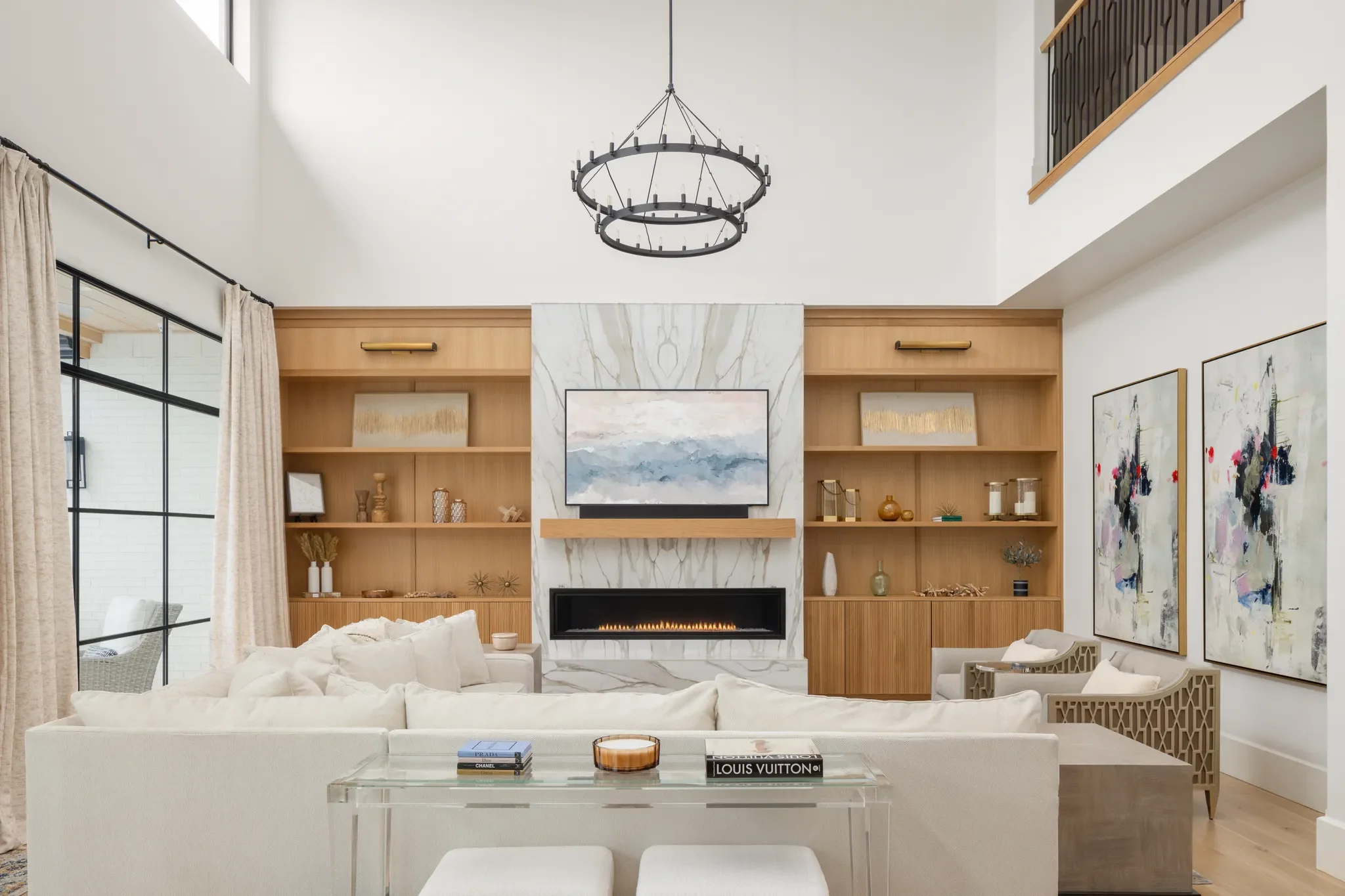 Modern living room with a white sofa, glass coffee table with books and decorative items, wooden shelves with art pieces, a fireplace with a marble surround, abstract paintings on the wall, large windows with curtains, and a black circular chandelier hanging from the ceiling.