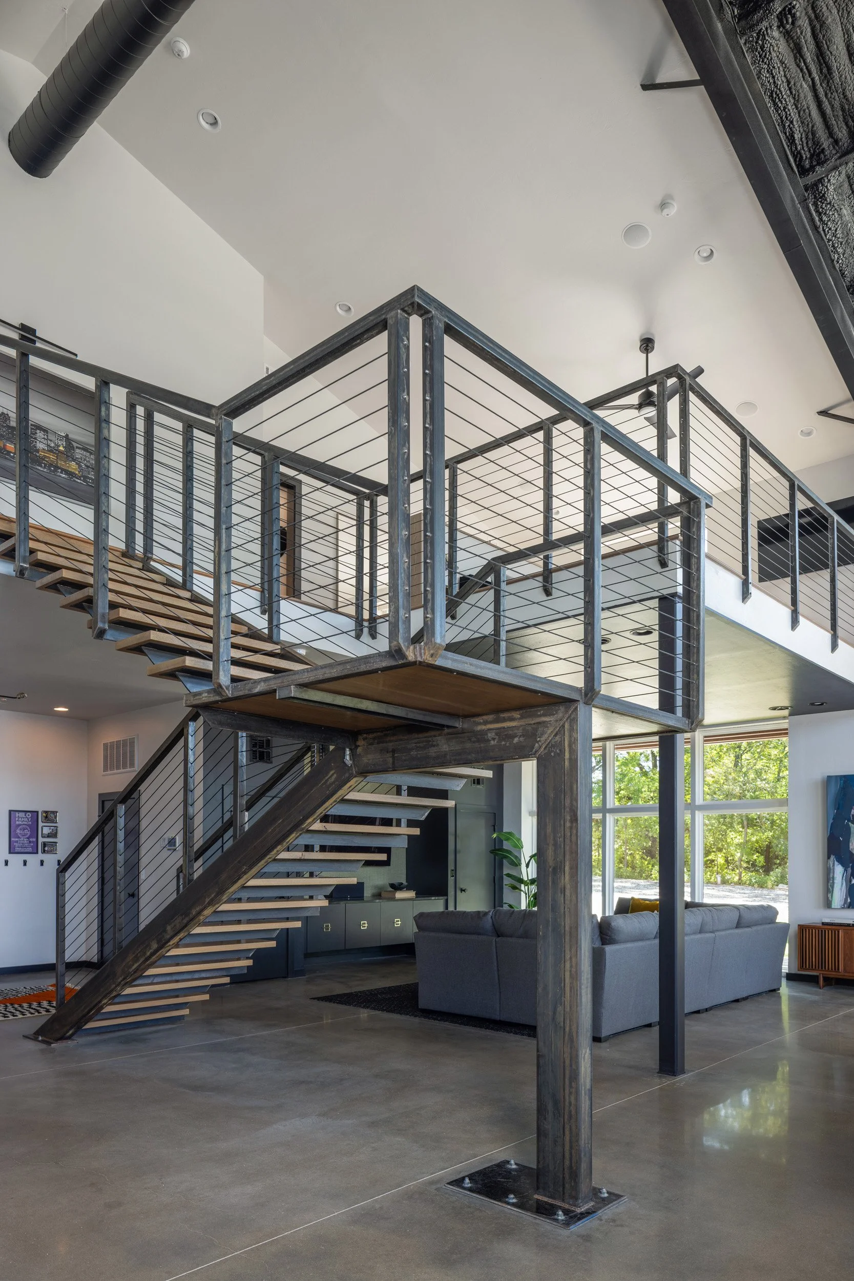 Modern interior with a staircase featuring metal railings and wooden steps, a gray sofa, large windows with a view of greenery outside, and a decorative plant near the windows.