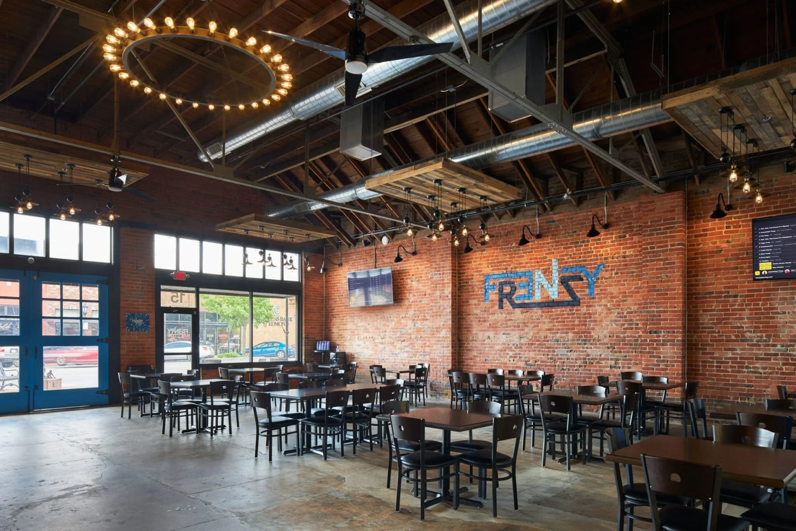Interior of a modern restaurant with exposed brick walls, industrial ceiling with ductwork, multiple black tables and chairs, two large digital screens, a decorative circular chandelier, and natural light from large windows.