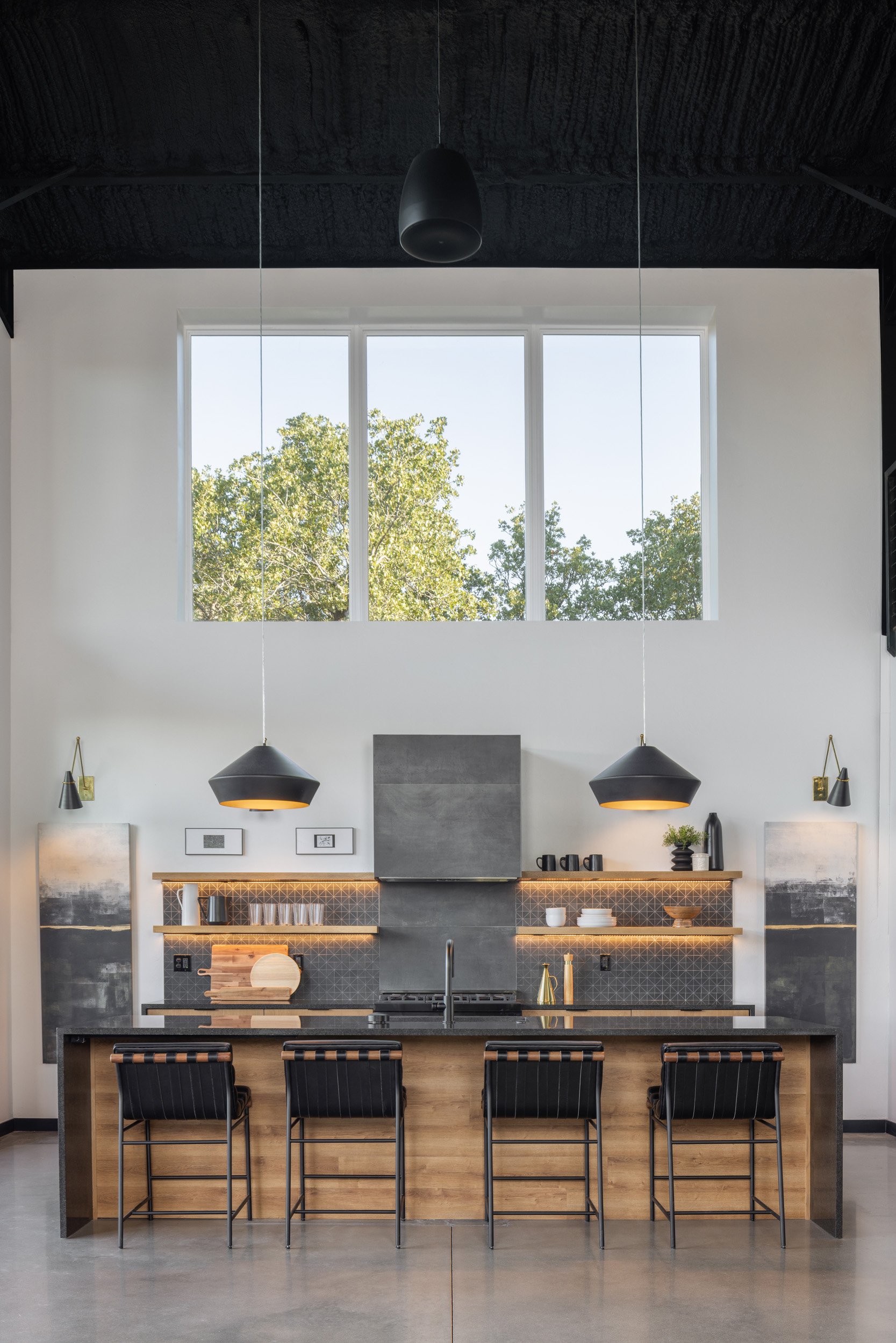 Modern kitchen interior with a large window, black pendant lights, and a wooden island with black chairs.