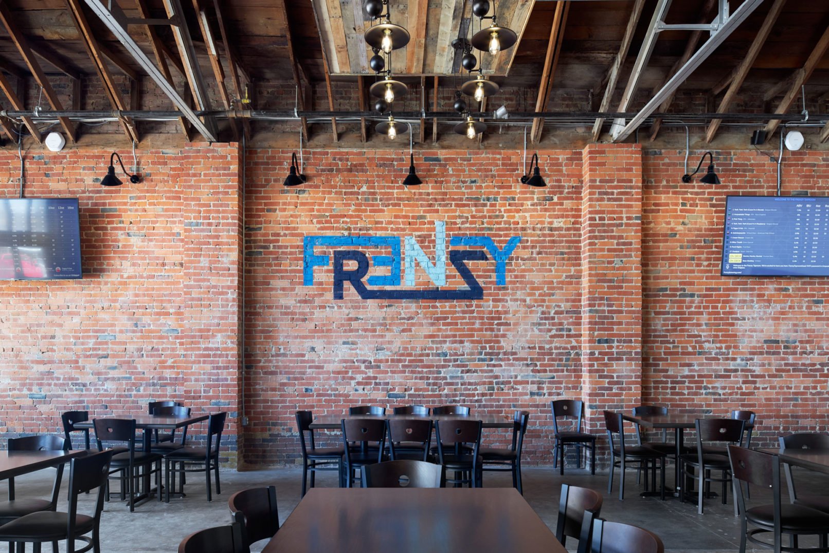 An inside view of a modern, industrial-style cafe with a brick wall featuring a large painted sign that says 'FRENZY'. The space has black chairs, tables, and mounted digital screens displaying information, with exposed wooden beams and hanging light
