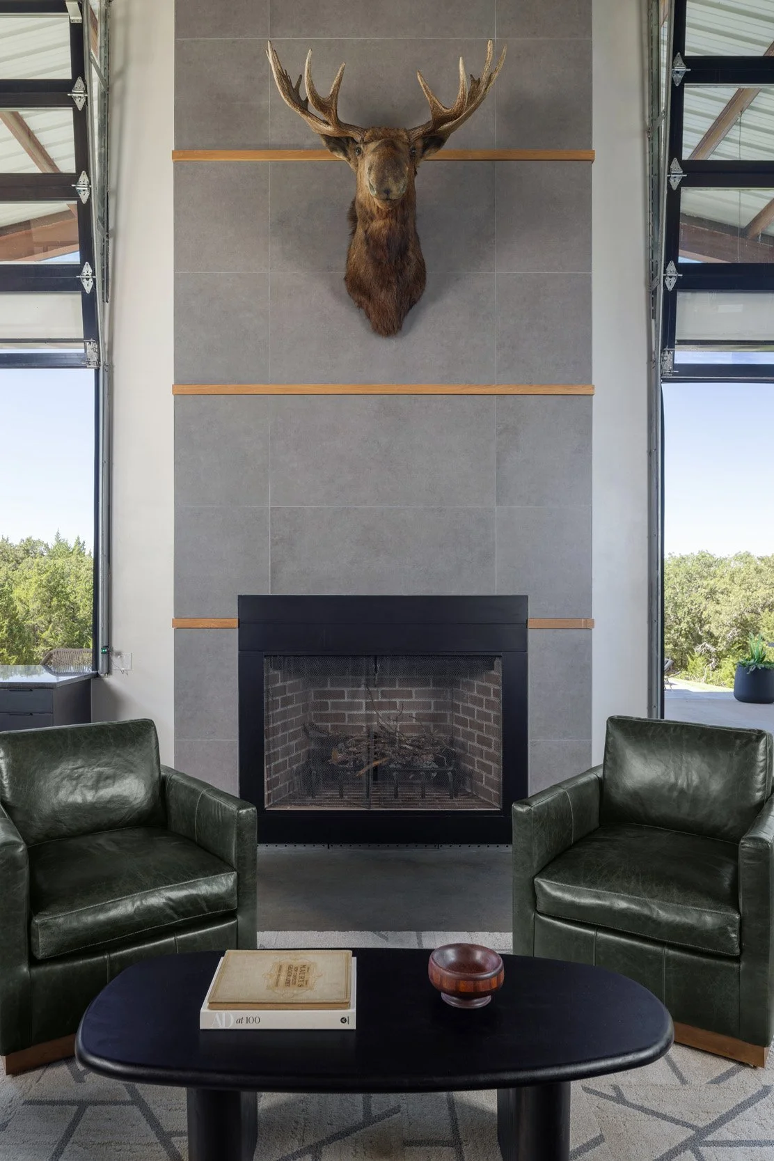 A modern living room with a tall gray tiled fireplace featuring a mounted deer head with large antlers. Two green leather armchairs are positioned on either side of a black oval coffee table, which holds a book and a small decorative bowl. Large wind
