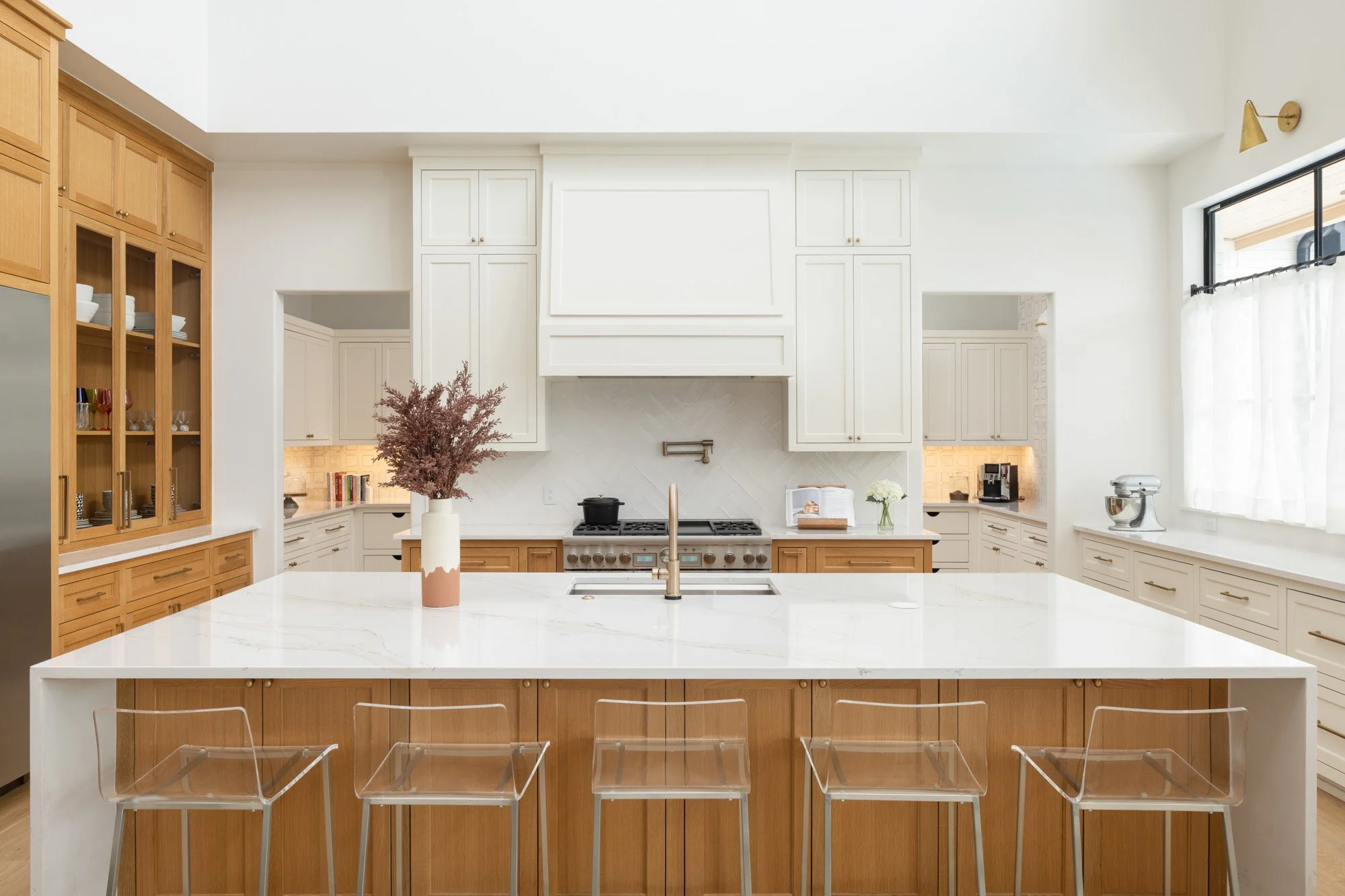 Modern kitchen with white walls, wooden cabinets, and a large white island in the center. There are clear acrylic chairs at the island, a vase with dried flowers, and small kitchen appliances on the countertops. Large windows with white curtains let in natural light.