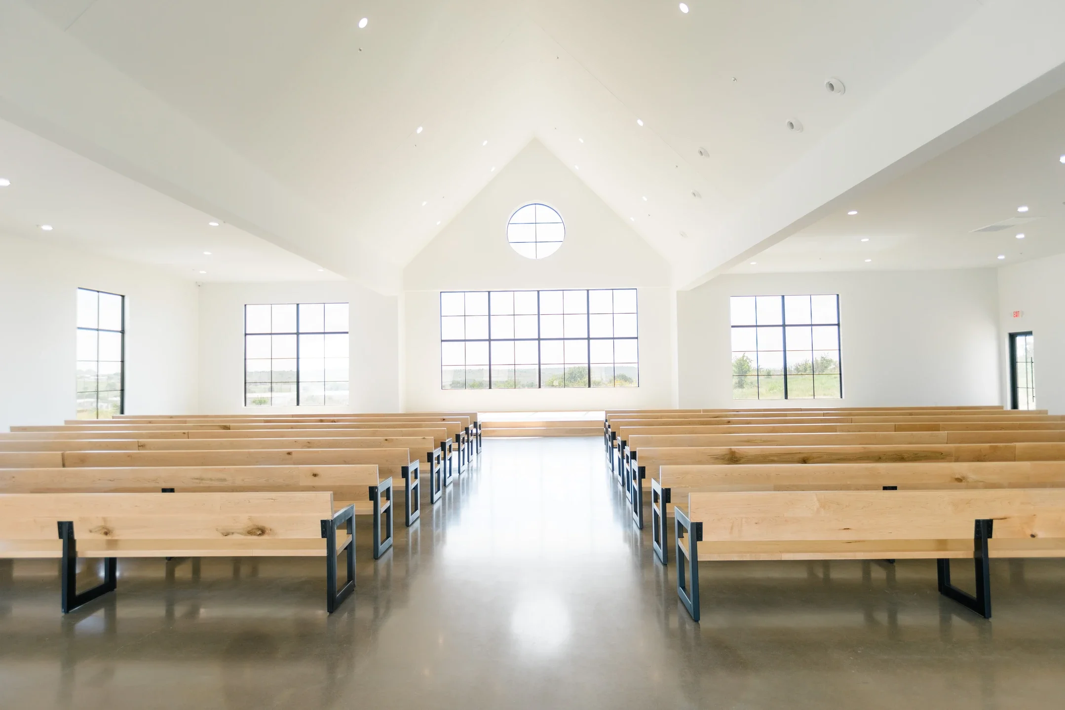 Interior of a modern church or chapel with rows of wooden pews, large windows, and a high vaulted ceiling with recessed lighting, bright with natural light.