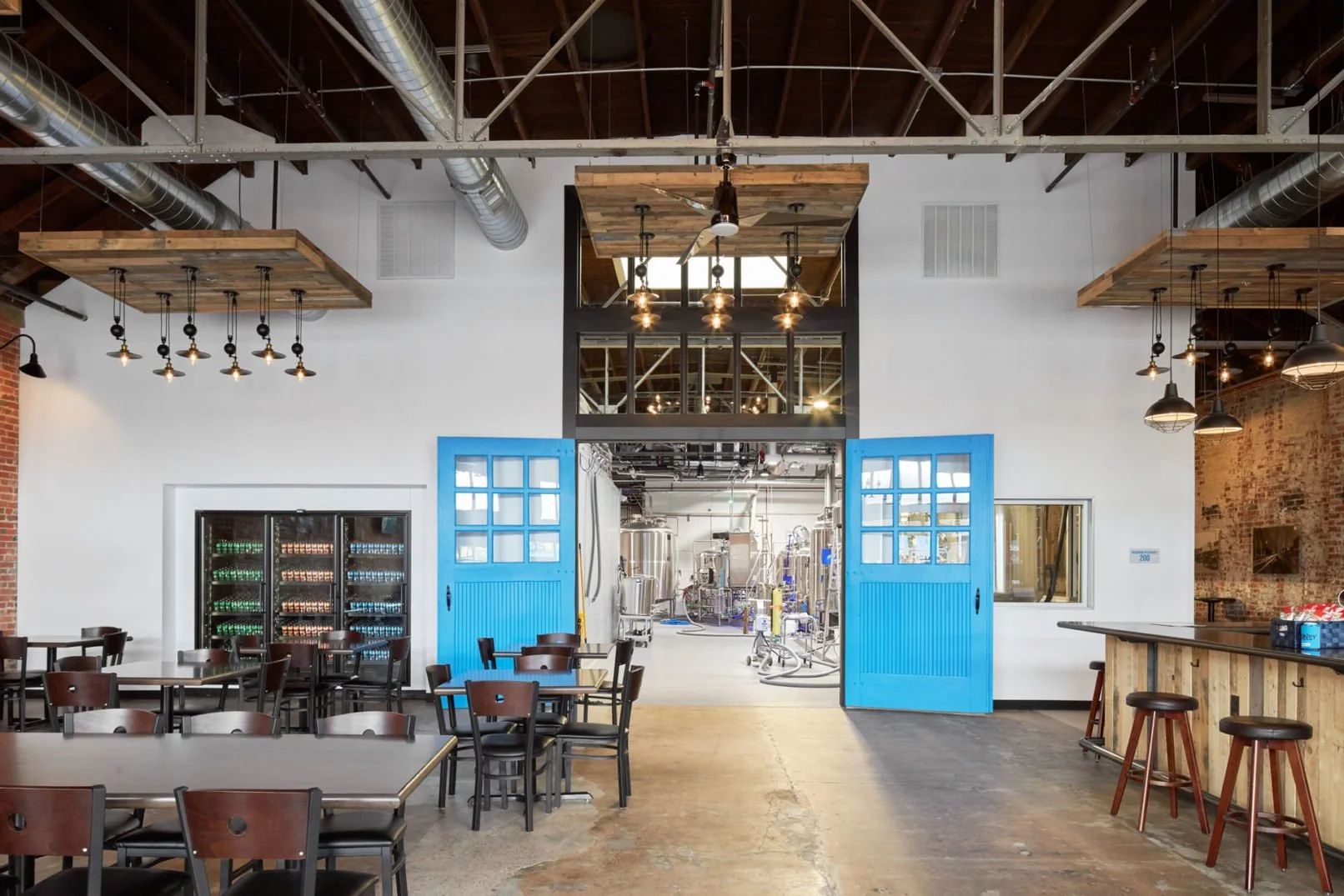Interior of a brewery or taproom with wooden and industrial design, blue sliding doors leading to brewing equipment, seating area with tables and chairs, and a bar counter on the right.