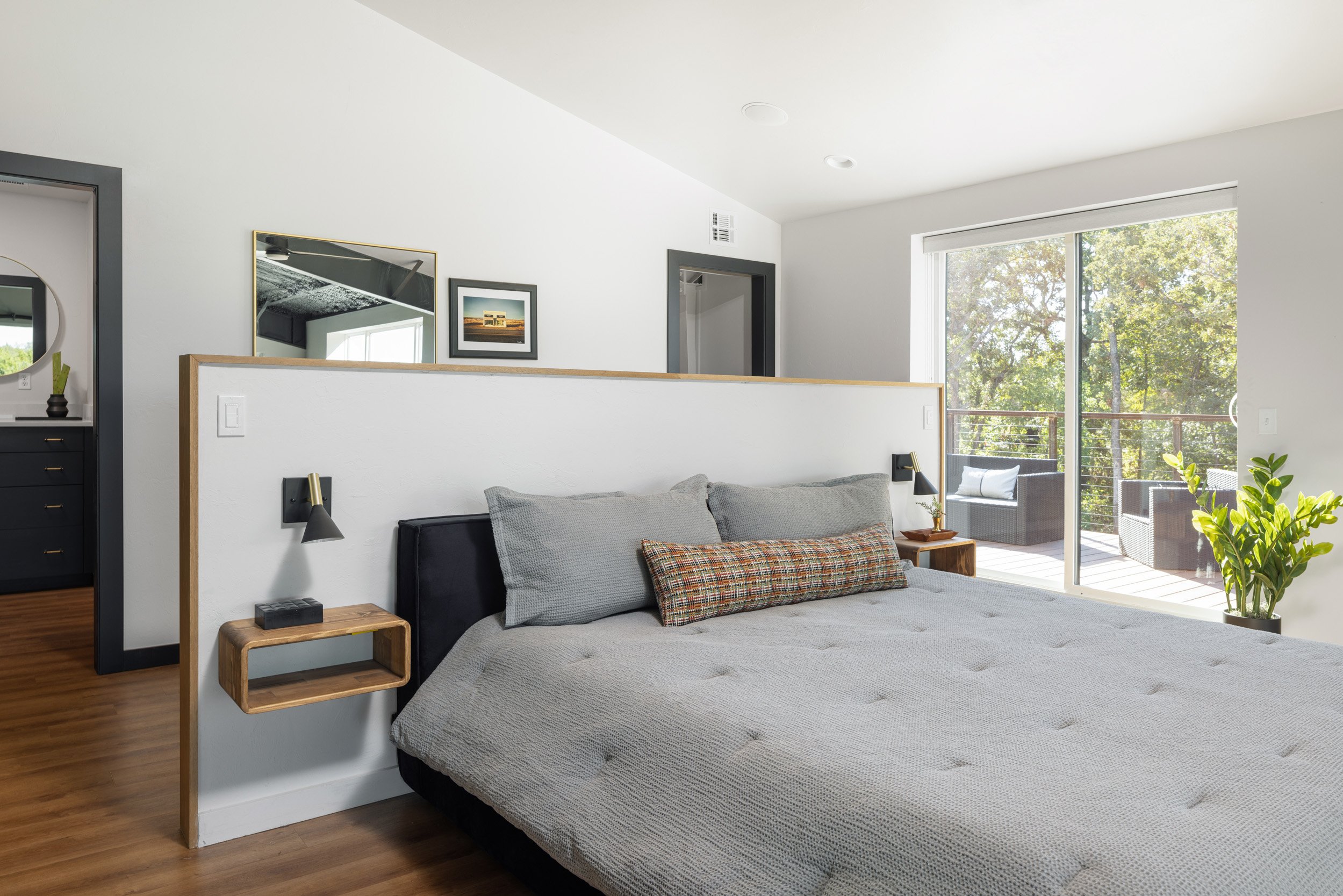 A bedroom with a large window showing trees outside, a bed with a gray quilt and pillows, a small wooden side table, and decor such as framed pictures and plants.