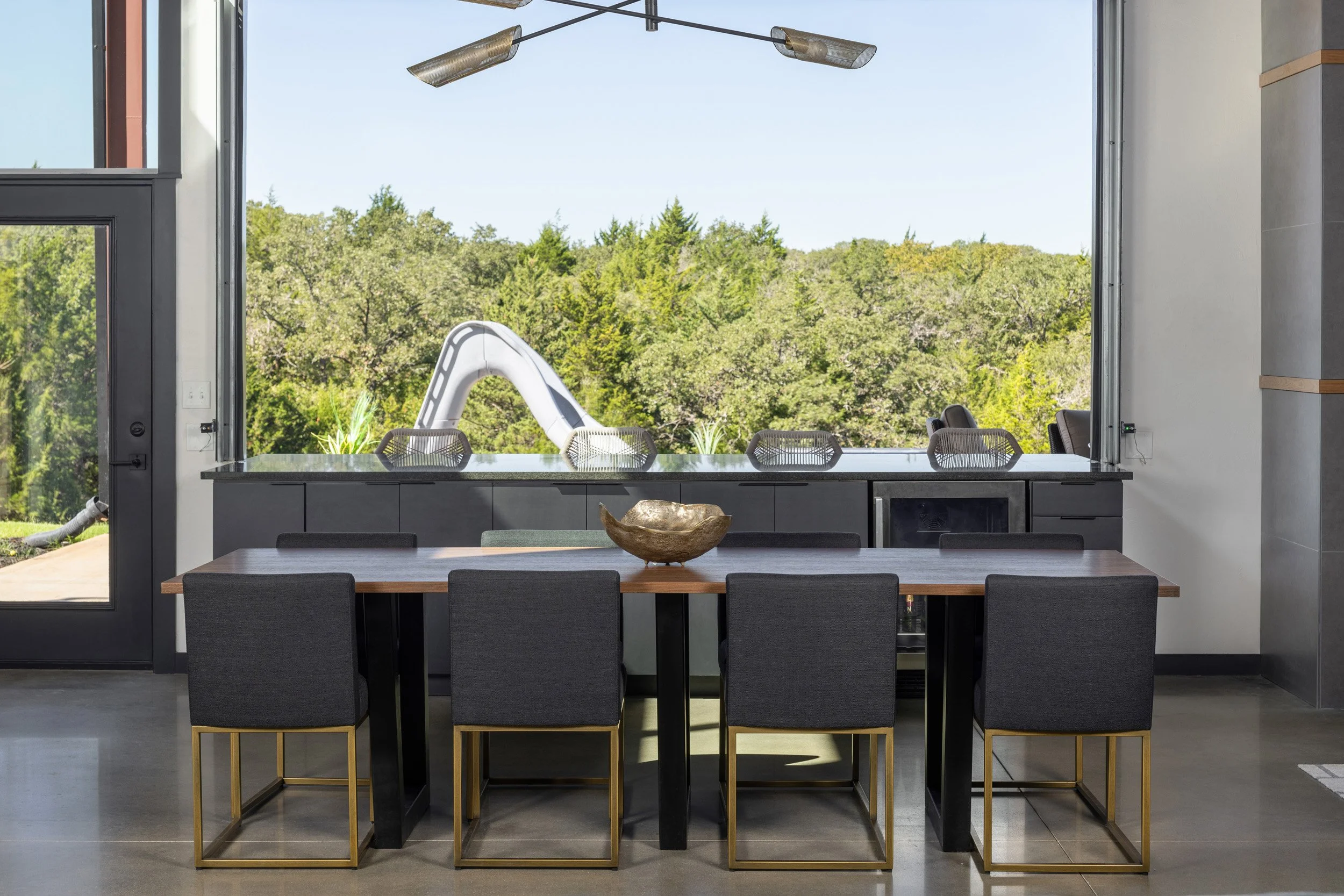 Modern indoor dining area with a large table, eight chairs, a decorative bowl on the table, and a scenic view of a lush green forest through open windows.