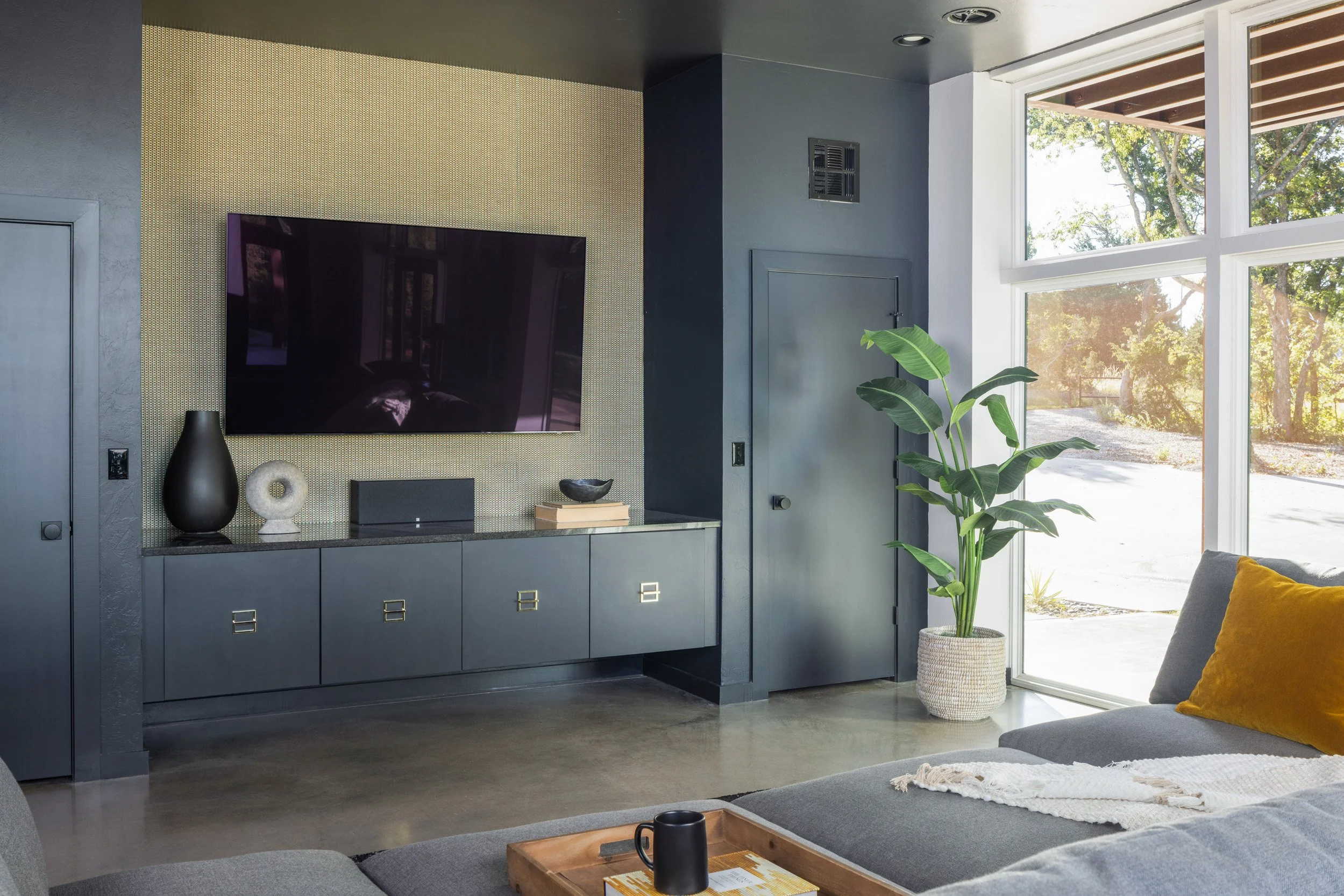 Living room with a gray and blue color scheme, large window showing trees outside, a flat-screen TV on the wall, decorative vases on a cabinet, a green potted plant, and a beige throw blanket on the sofa.