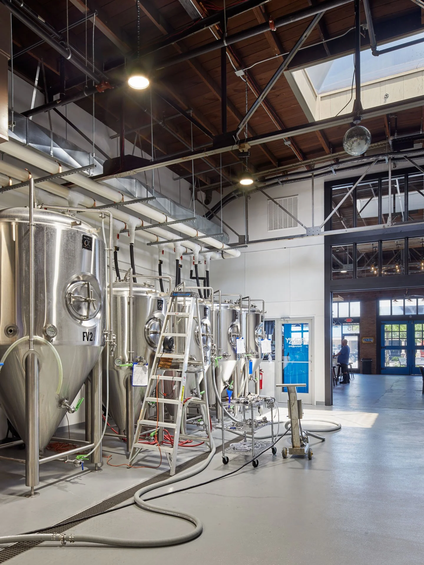 Interior of a brewery with large stainless steel fermentation tanks, pipes, and brewing equipment, lit by ceiling lights with a partial view of the outside through large windows.