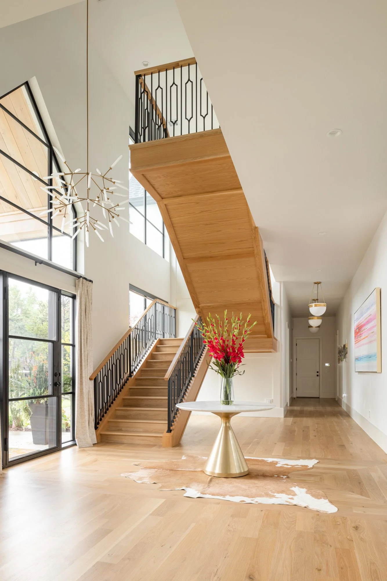 Modern interior featuring a staircase with wooden steps, black metal railings, large windows, a white ceiling, a chandelier, a white table with a gold base, a cowhide rug, and vibrant pink flowers in a glass vase.