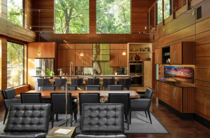 Modern kitchen and dining area with wooden walls, large windows, and a high ceiling, featuring a long dining table with black chairs, kitchen cabinets, and a television.