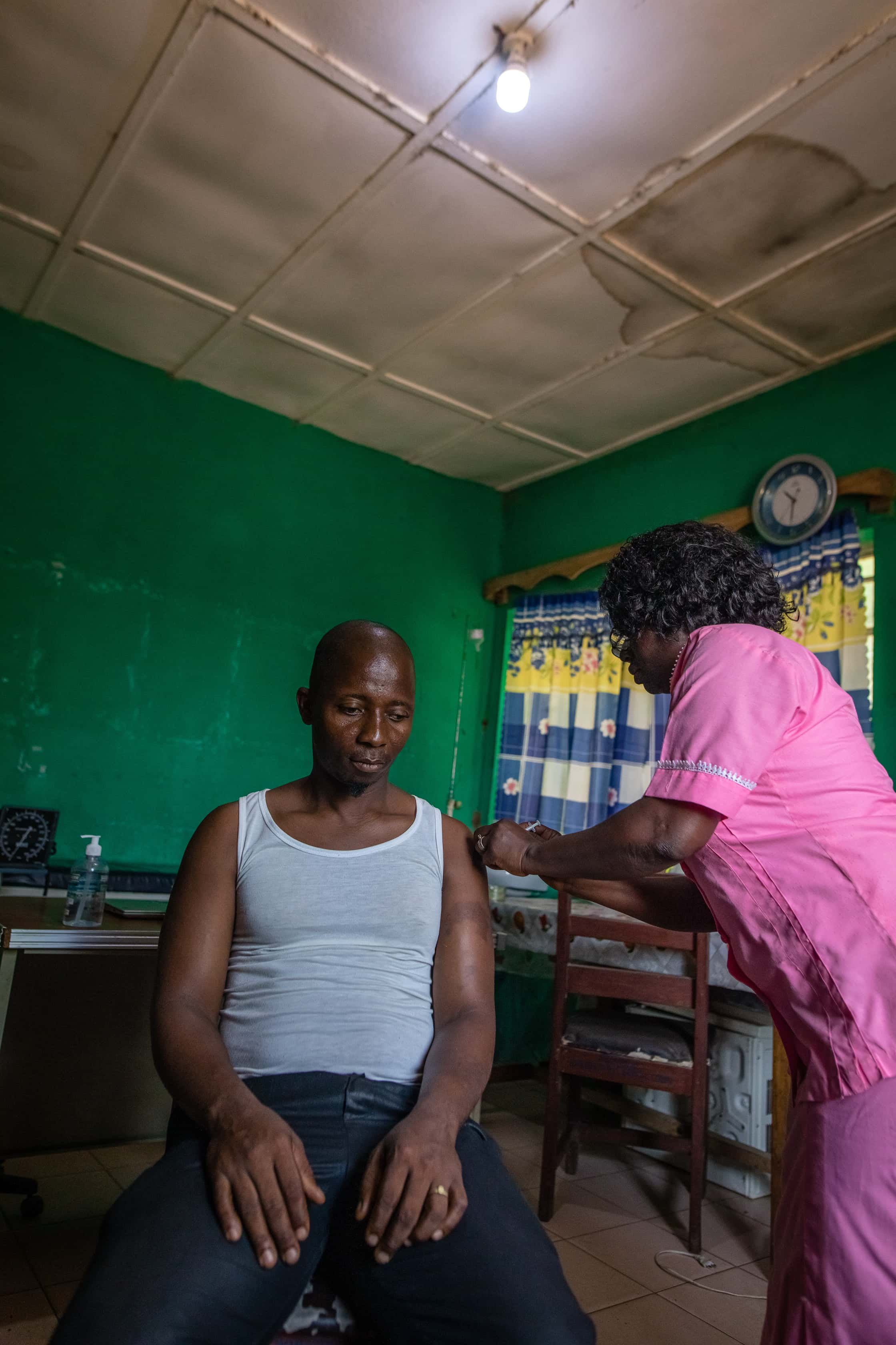 A man receiving a vaccination shot from a healthcare worker in a room with green walls, a clock, and curtains.