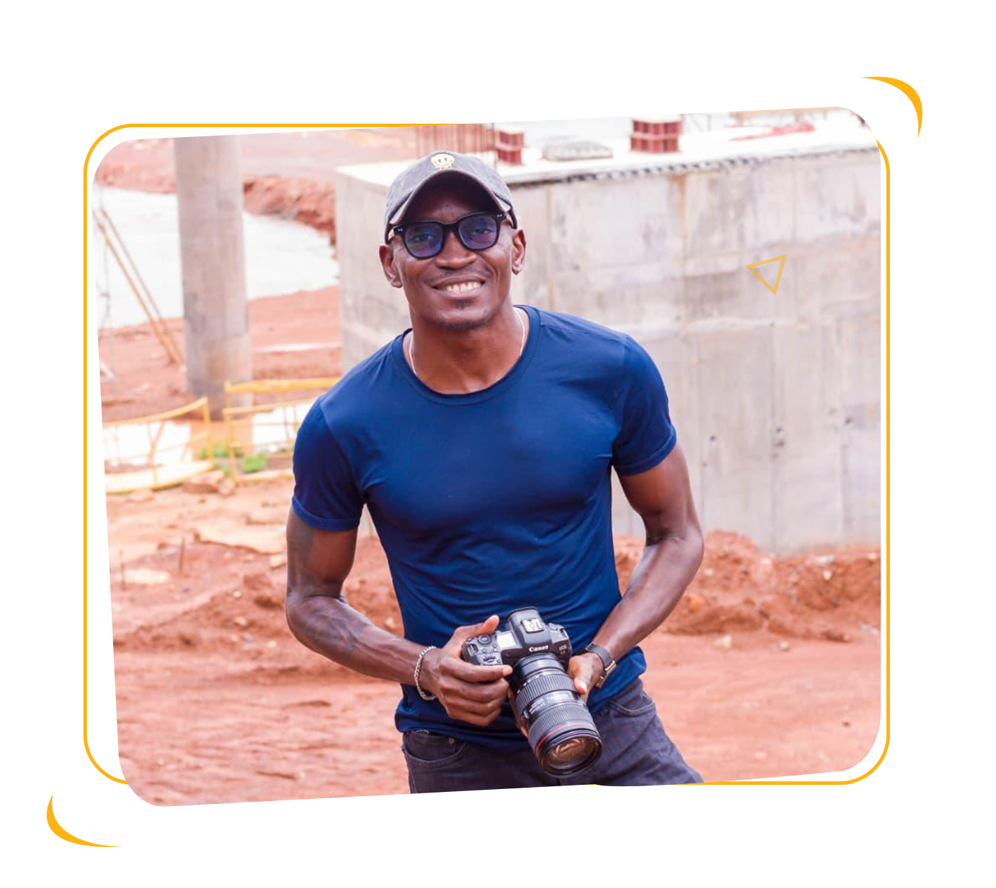A smiling man wearing sunglasses, a black cap, and a blue t-shirt, holding a professional camera at a construction site with exposed soil and concrete structures.