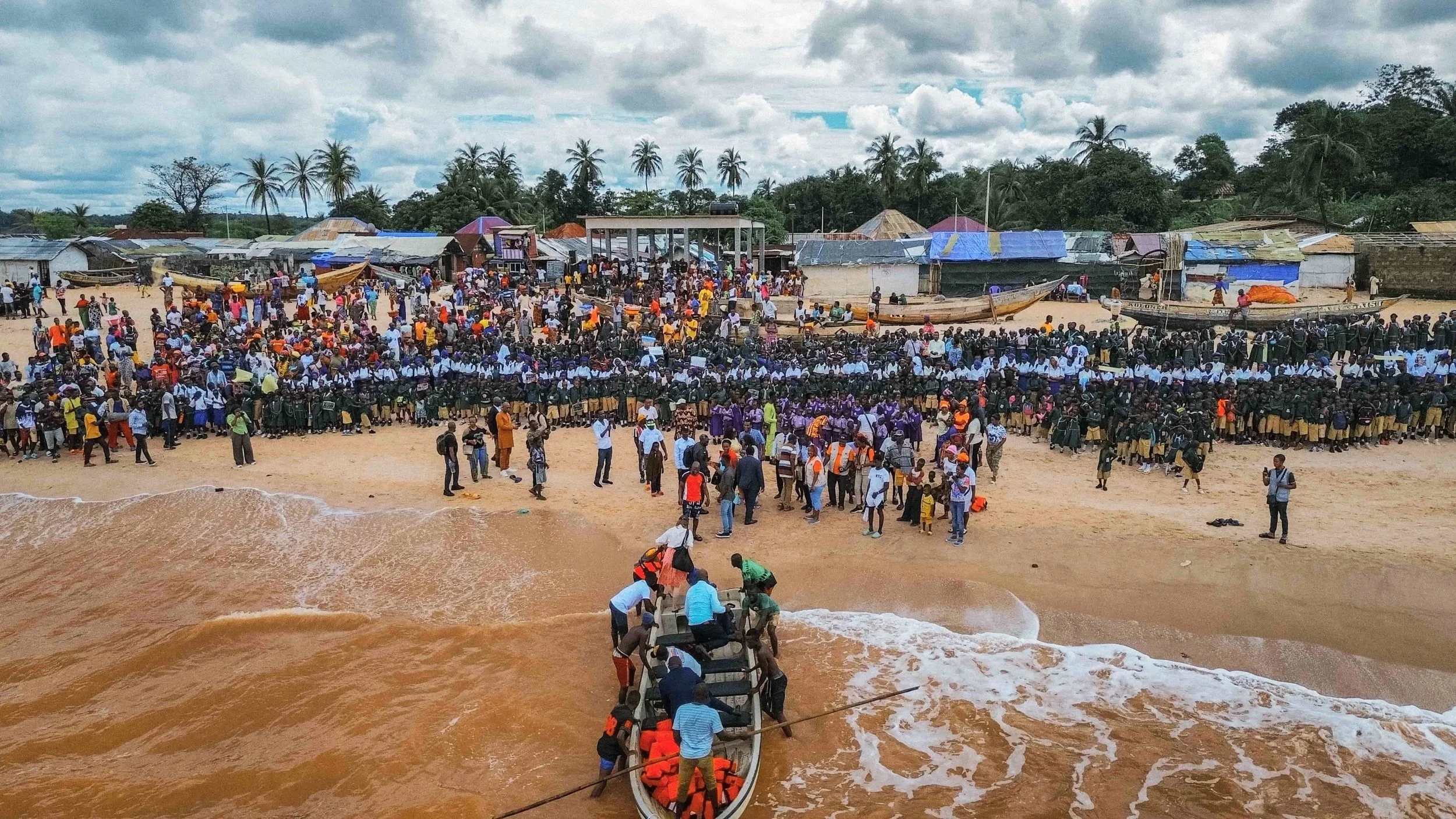 Large crowd of people gathered on a beach with boats and houses in the background. Some people are entering the water by boat, with others watching or walking along the shore.