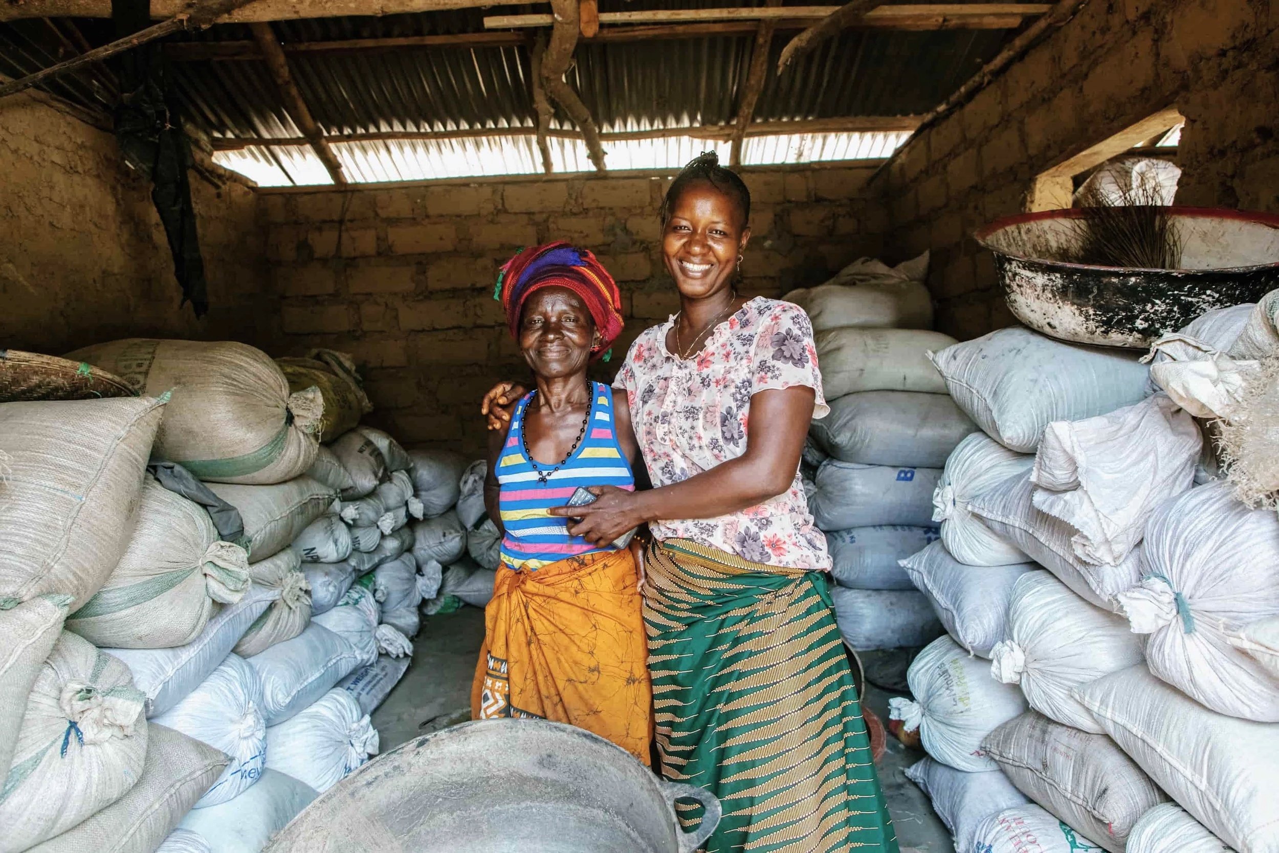 Two women standing inside a storage room filled with large bags of materials, smiling and posing for the camera.