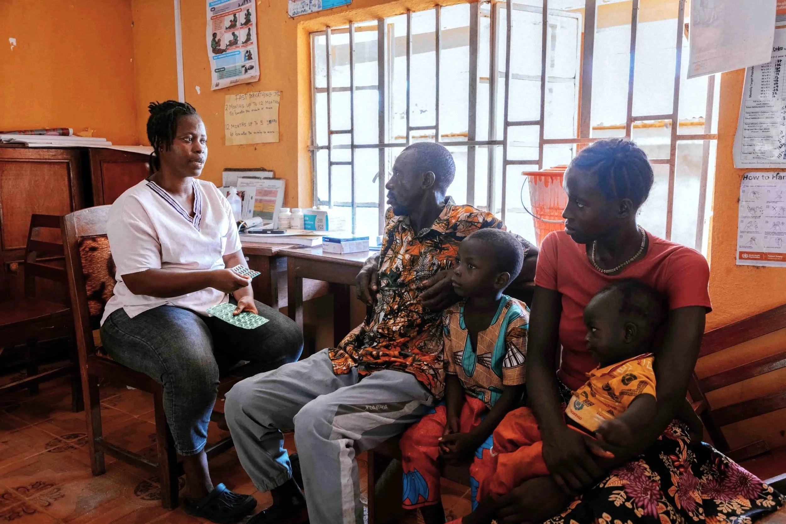 A healthcare professional sitting on a wooden chair, talking to a group of four people, including children and adults, inside a room with orange walls and posters on the walls. There is a window with bars behind them.