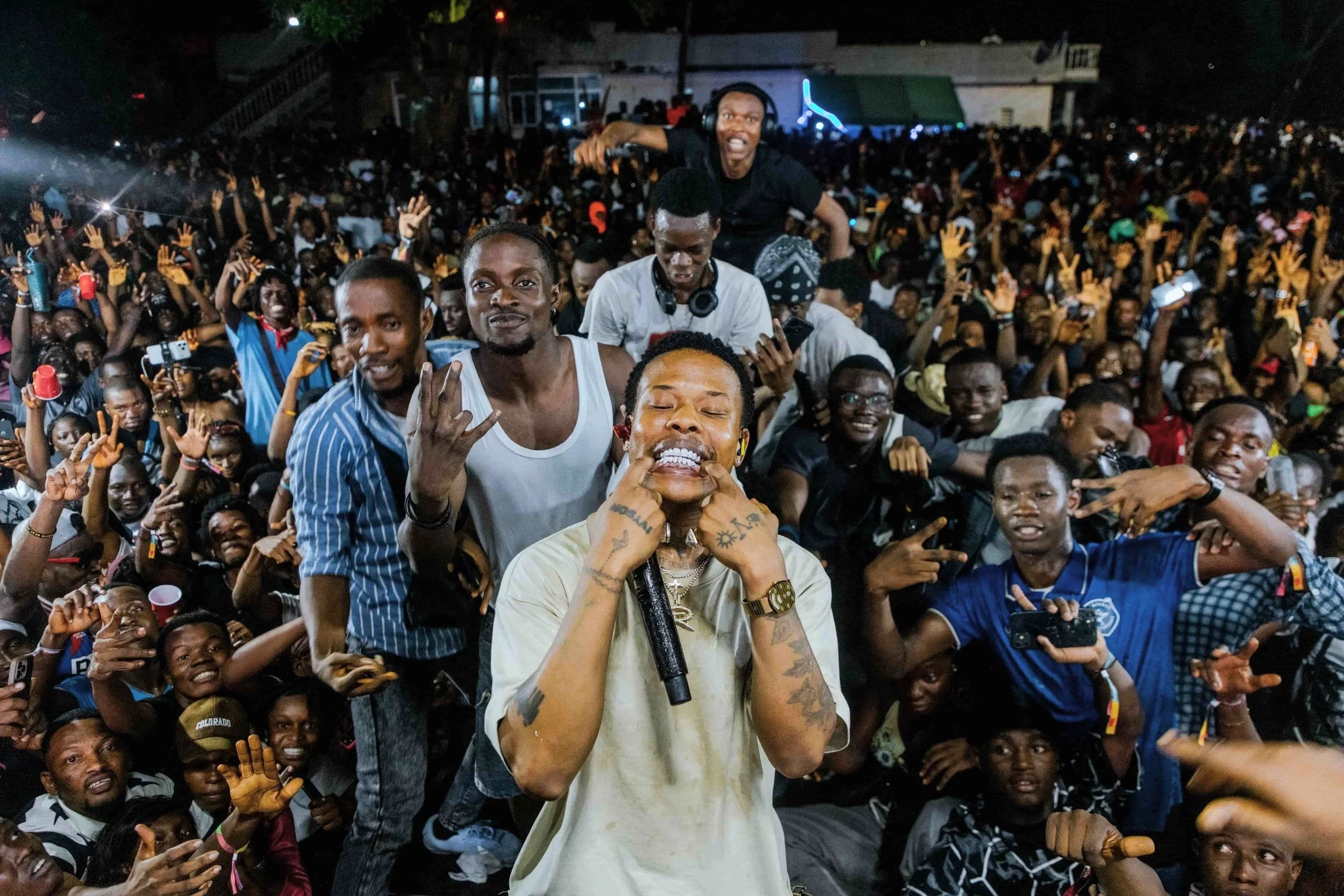 A large crowd of people at night, with some holding up their hands and taking photos, and a person in the foreground holding a microphone and making a peace sign.
