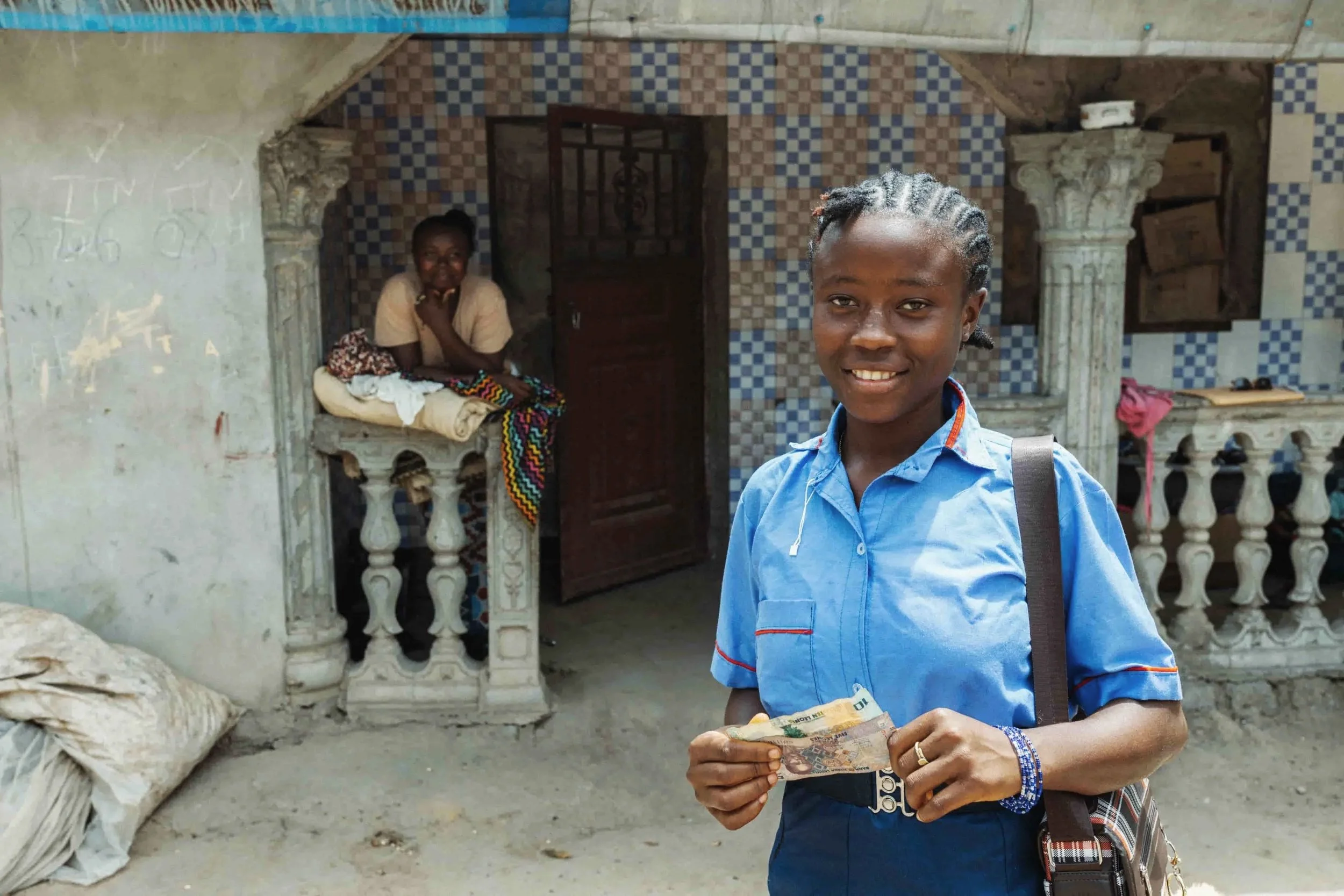 A young person in a blue shirt holding money in front of an ornate building with two women sitting on a balcony behind him.