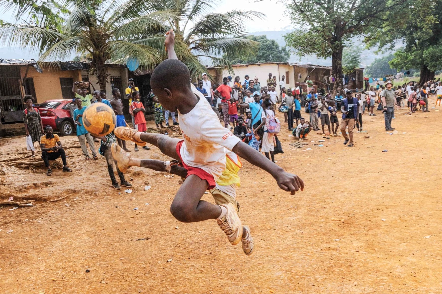 A boy is jumping in the foreground while playing with a ball. Behind him, a large crowd of people, including children and adults, are gathered outdoors in a rural area. There are houses, trees, and vehicles visible in the background.