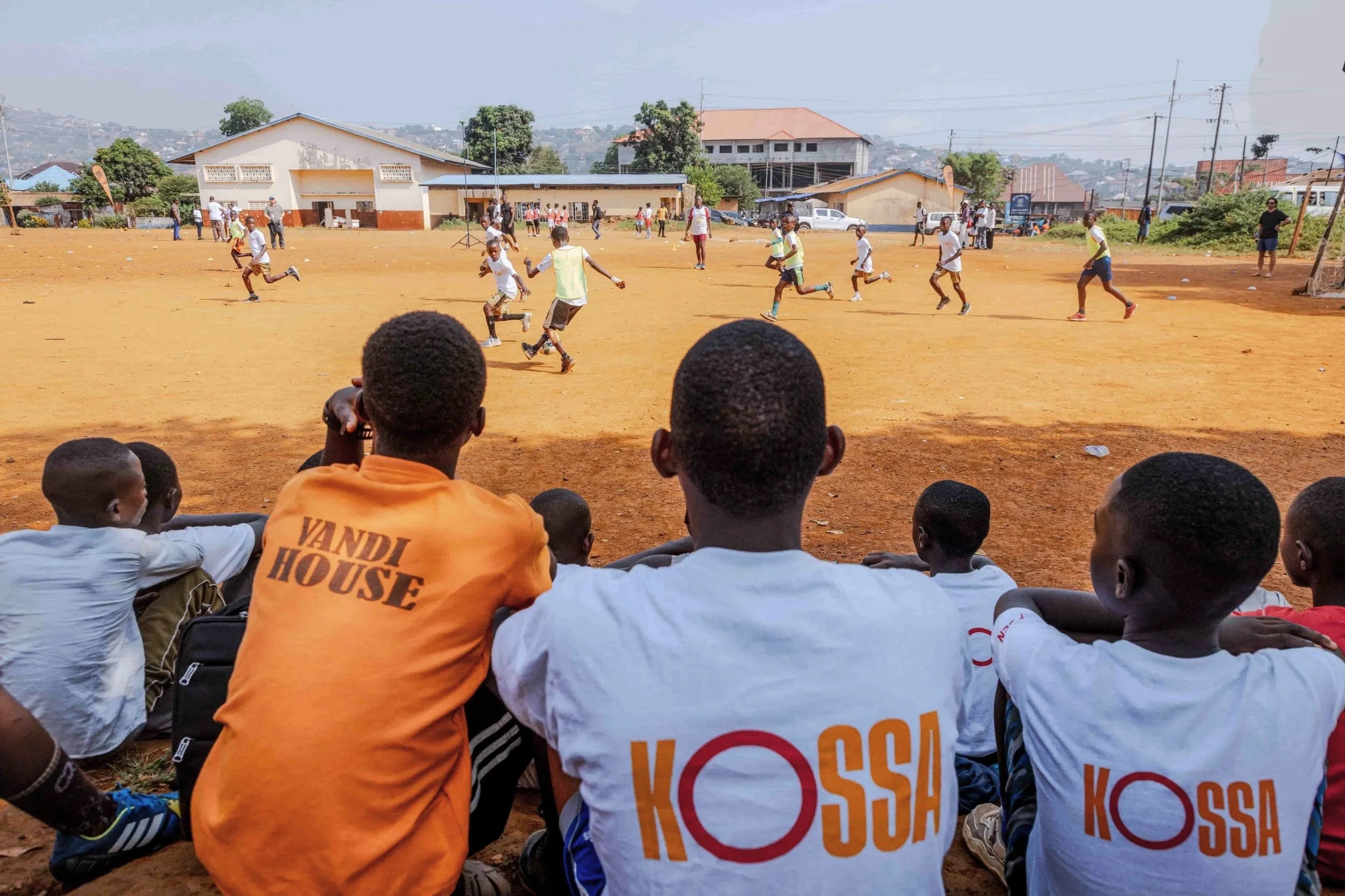 Children playing soccer on a dirt field, with spectators sitting at the edge of the field, in a community with buildings and trees in the background.