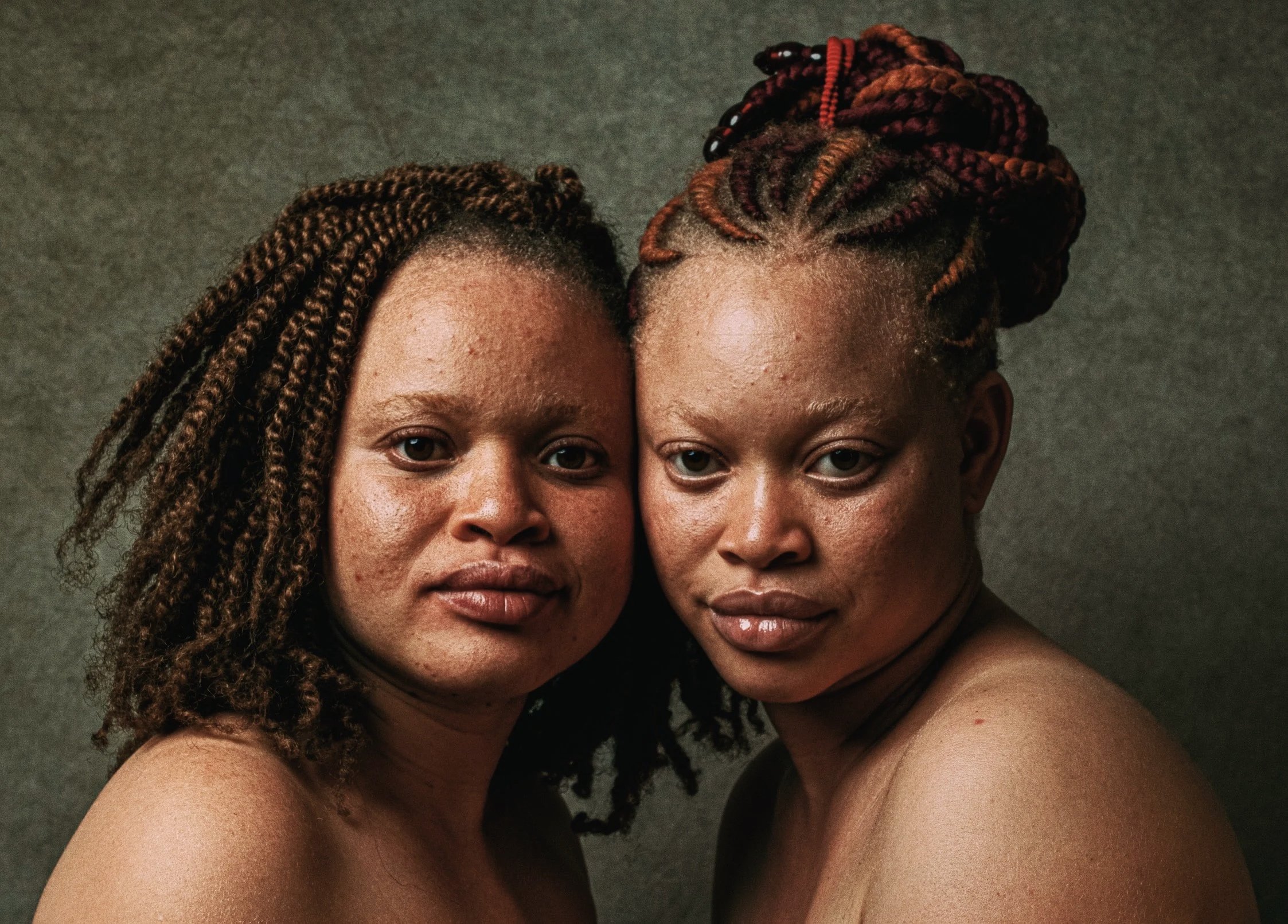 Close-up of two women with their faces close together, showing their shoulders and part of their arms, with brown and reddish braided hairstyles, set against a gray background.