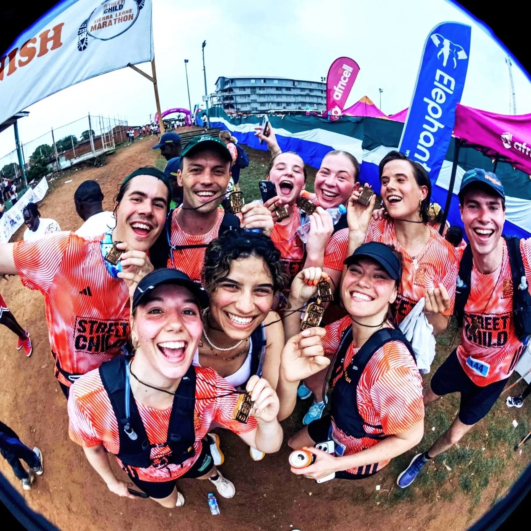Group of smiling marathon finishers in orange and red shirts holding medals at a race event.