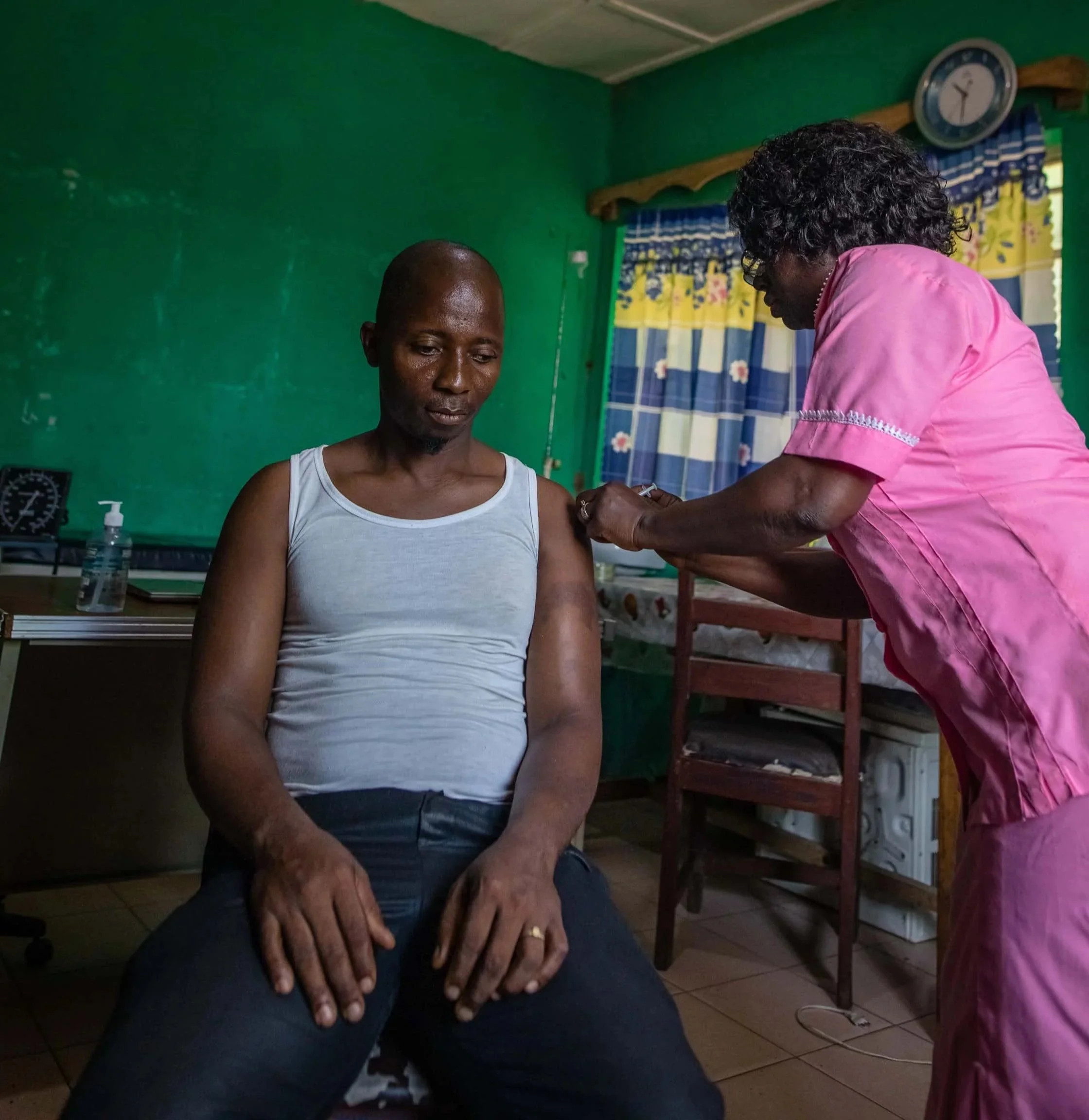 A man receiving a vaccination shot from a healthcare worker in a room with green walls, a clock, and curtains.