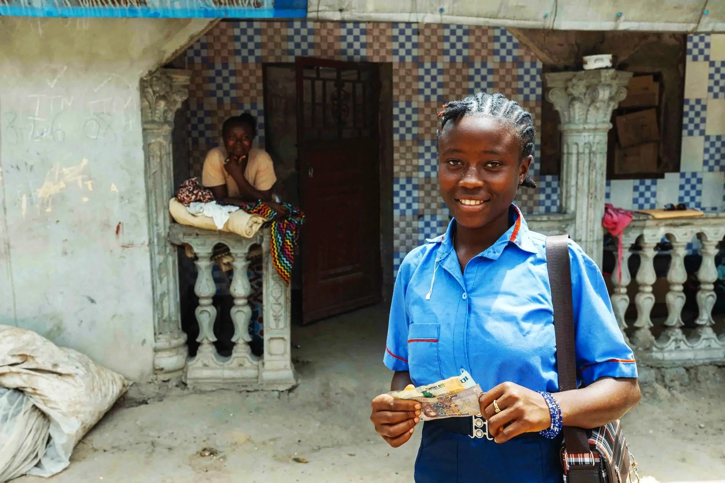 A young person in a blue shirt holding money in front of an ornate building with two women sitting on a balcony behind him.