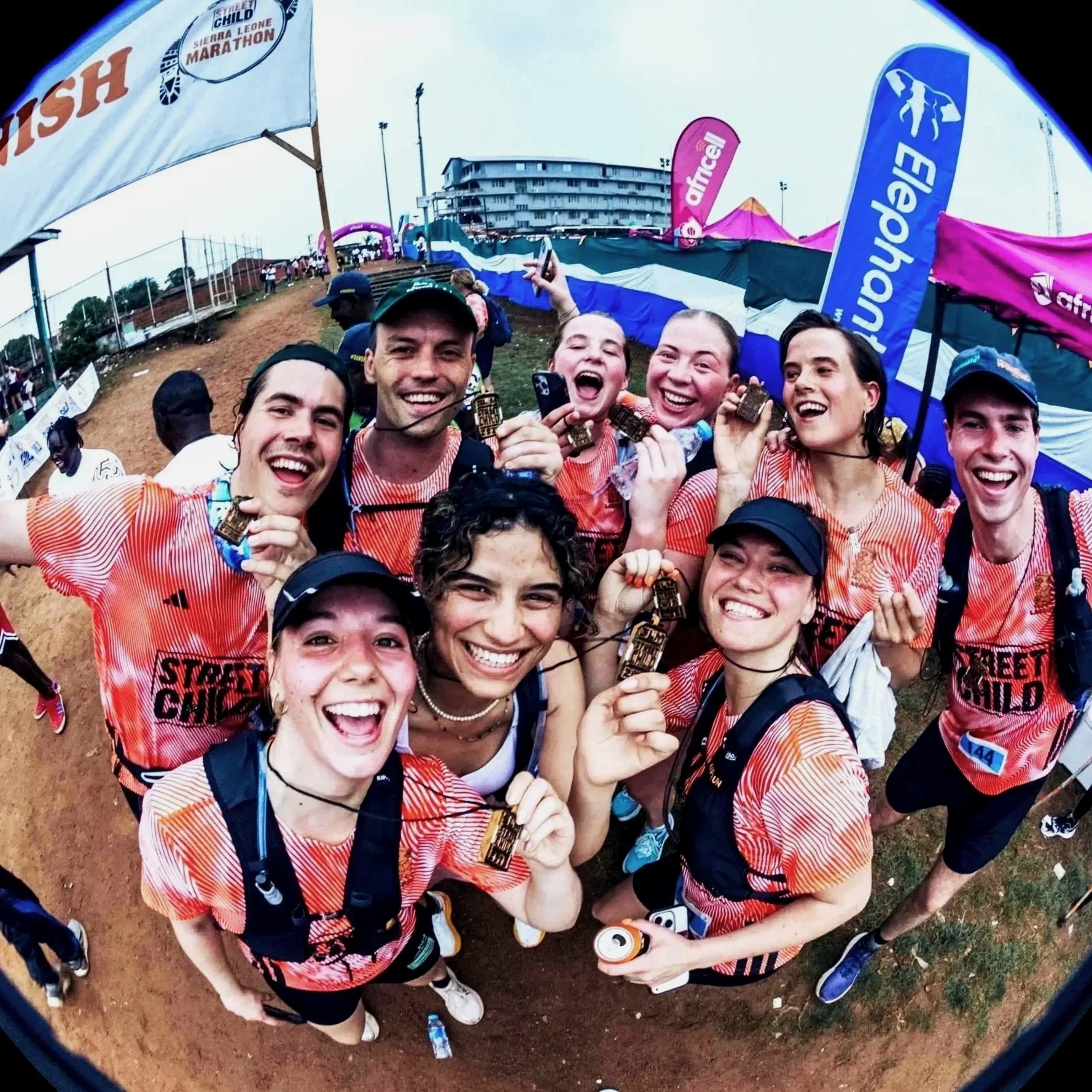 Group of smiling marathon finishers in orange and red shirts holding medals at a race event.