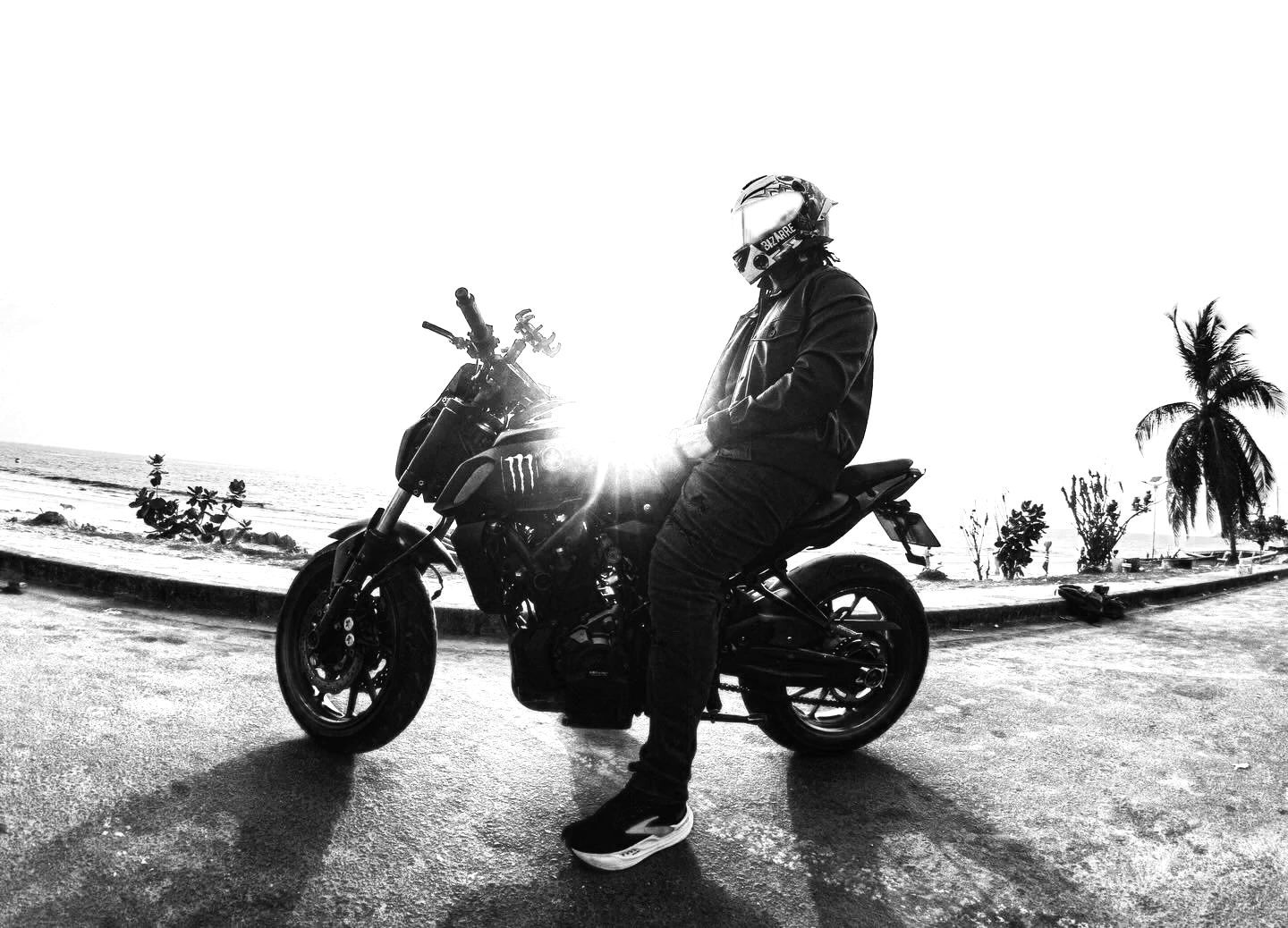 Person wearing a helmet and black riding gear sitting on a black motorcycle near the beach during sunset with palm trees in the background.